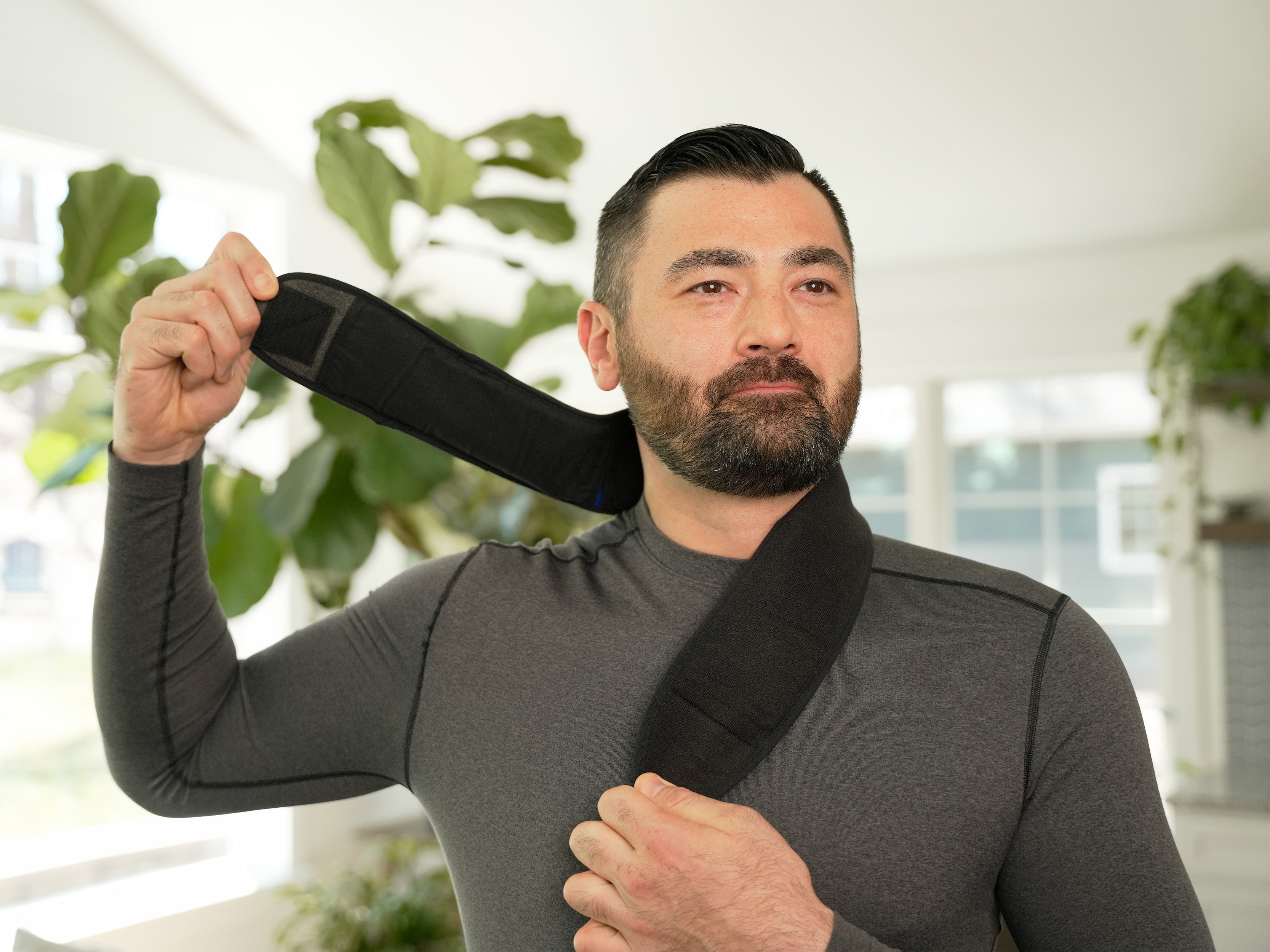 Man applying a Soft Ice® Neck & Upper Spine Wrap in a living room setting with plants in the background.