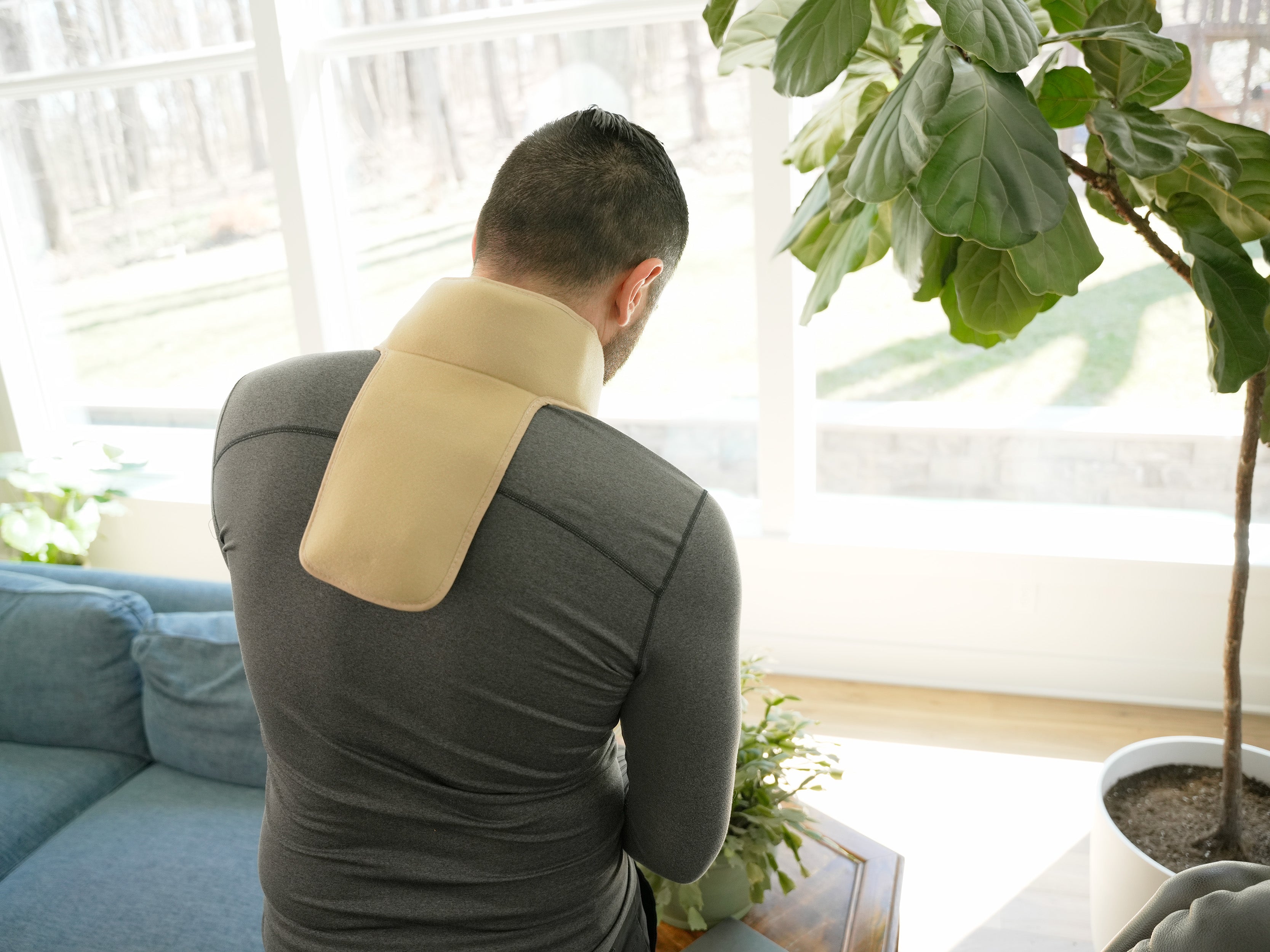 Man watering plants wearing a khaki Soft Ice® Neck & Upper Spine Wrap in a living room setting.
