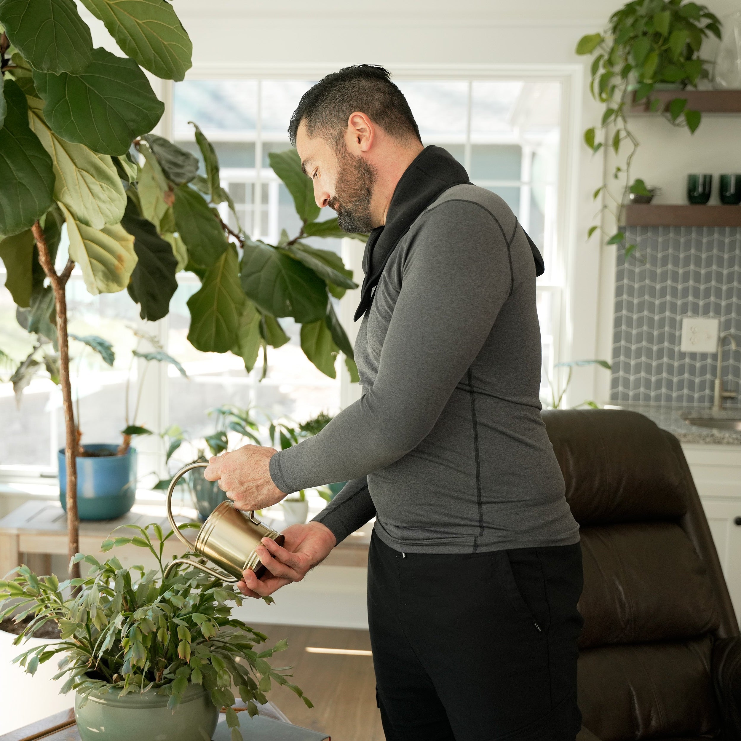 Man watering plants wearing a black Soft Ice® Neck & Upper Spine Wrap in a living room.