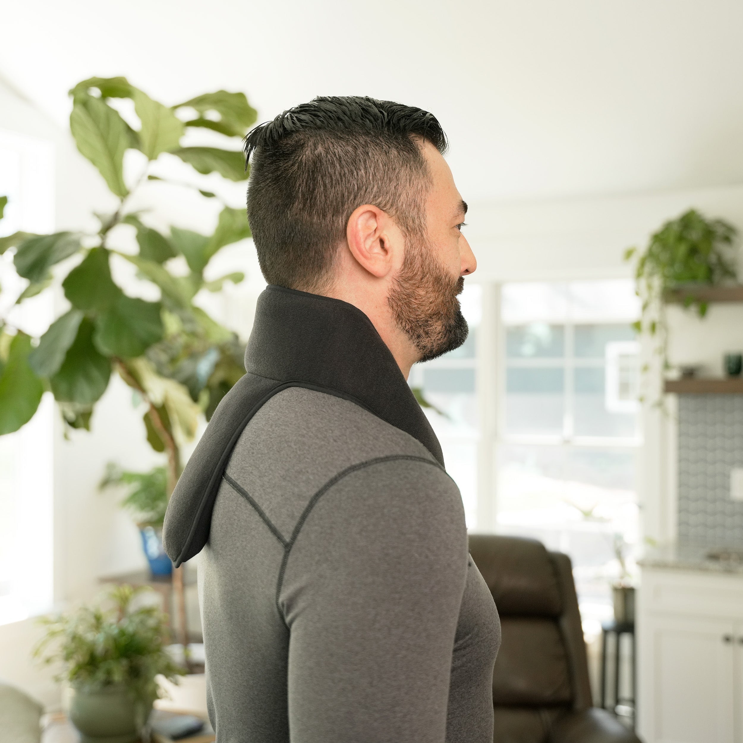 Man wearing a gray long sleeve wearing a black Soft Ice® Neck & Upper Spine Wrap in a living room setting.