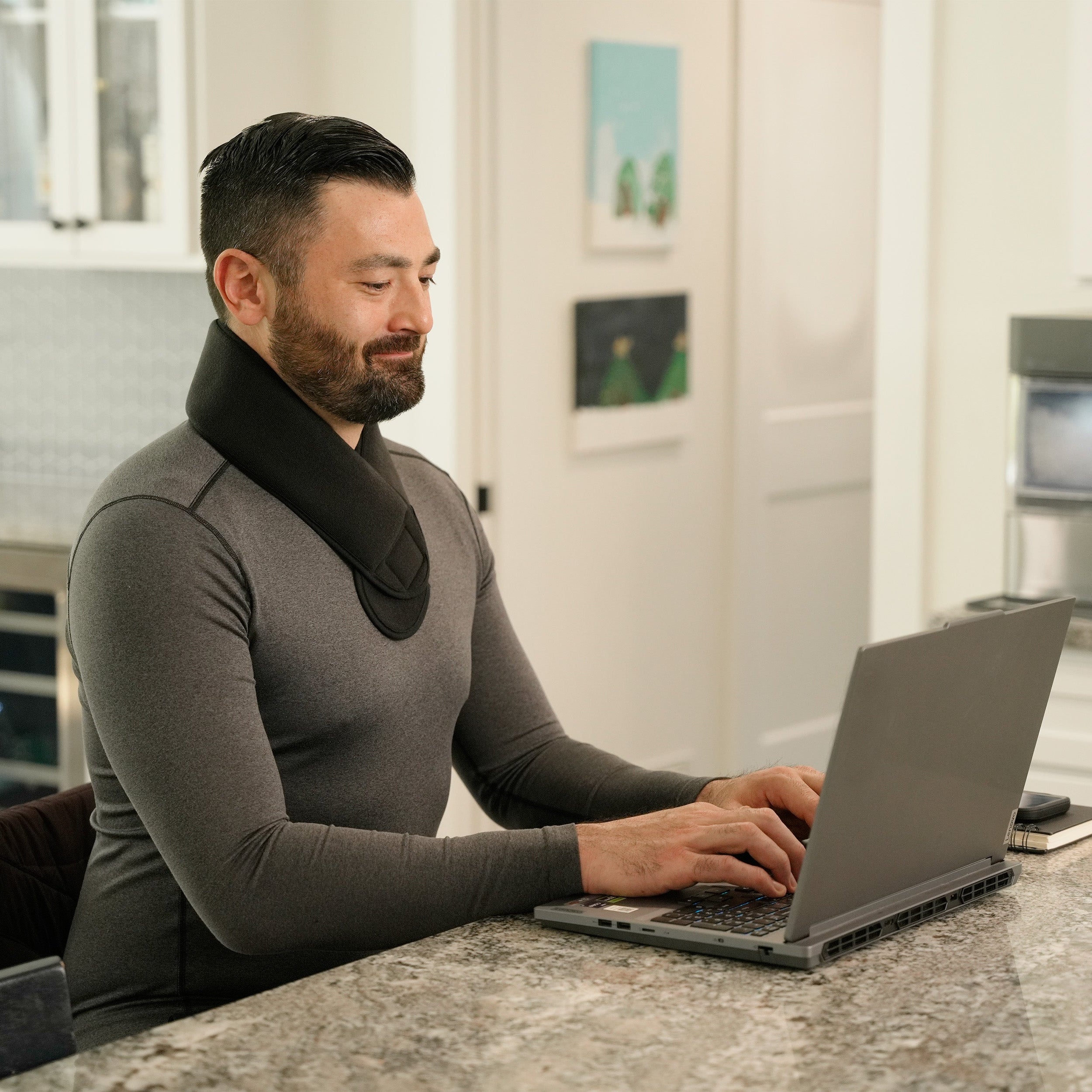 Man wearing a Soft Ice® Neck and Head Wrap in a sitting at the kitchen island working on a laptop.