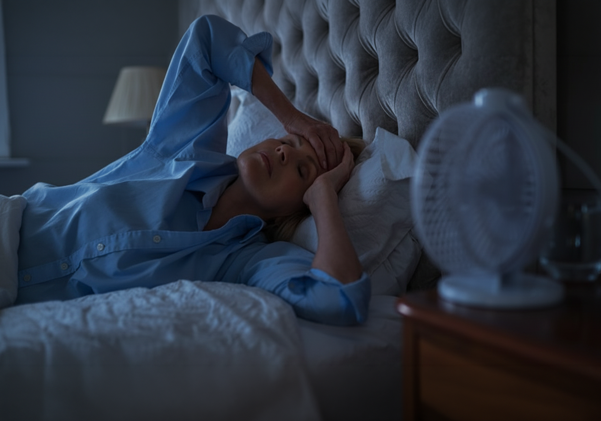 A woman in blue pajamas lies in bed with her head on a pillow and hands over her face, in a dimly lit bedroom with a bedside fan on the table.