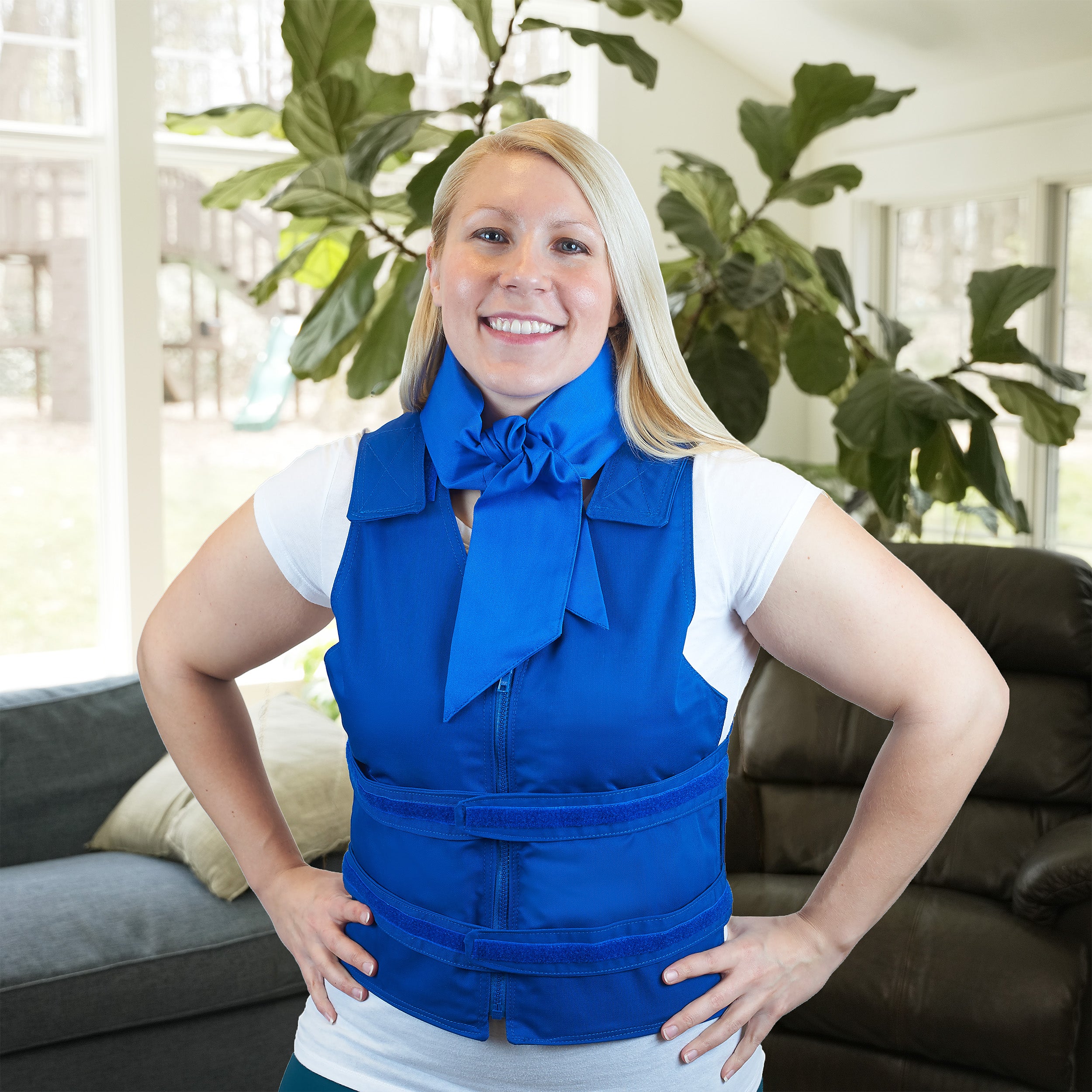 A smiling blonde woman wearing a bright blue deluxe cooling neck wrap and matching blue adjustable cooling vest with a front zipper, standing indoors in a living room with a couch and large green plant in the background.