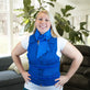 A smiling blonde woman wearing a bright blue deluxe cooling neck wrap and matching blue adjustable cooling vest with a front zipper, standing indoors in a living room with a couch and large green plant in the background.
