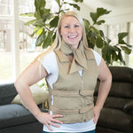 A smiling blonde woman wearing a beige adjustable cooling vest over a white t-shirt, with a khaki deluxe cooling neck wrap, standing in a living room with a couch and large green plants in the background.