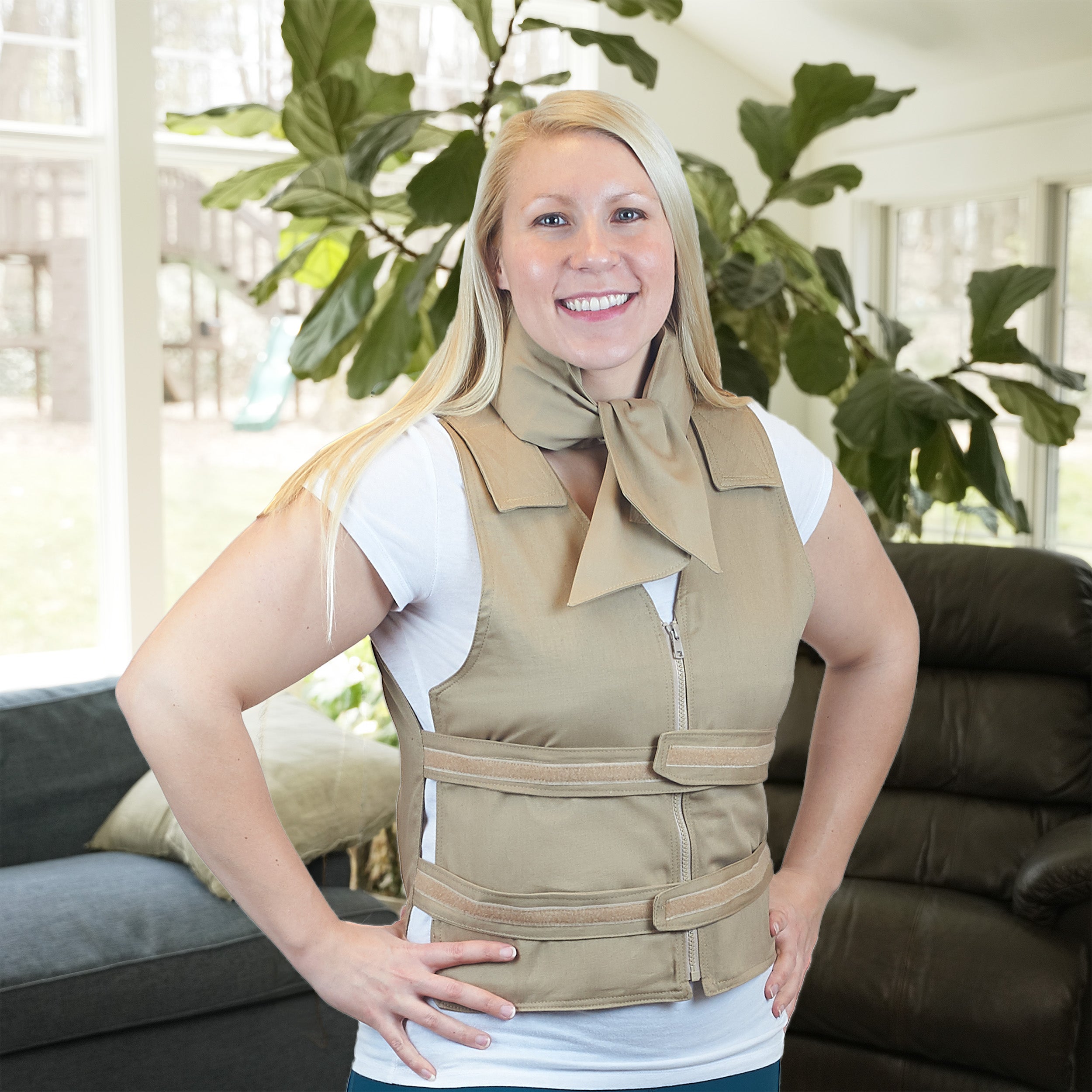 A smiling blonde woman wearing a beige adjustable cooling vest over a white t-shirt, with a khaki deluxe cooling neck wrap, standing in a living room with a couch and large green plants in the background.