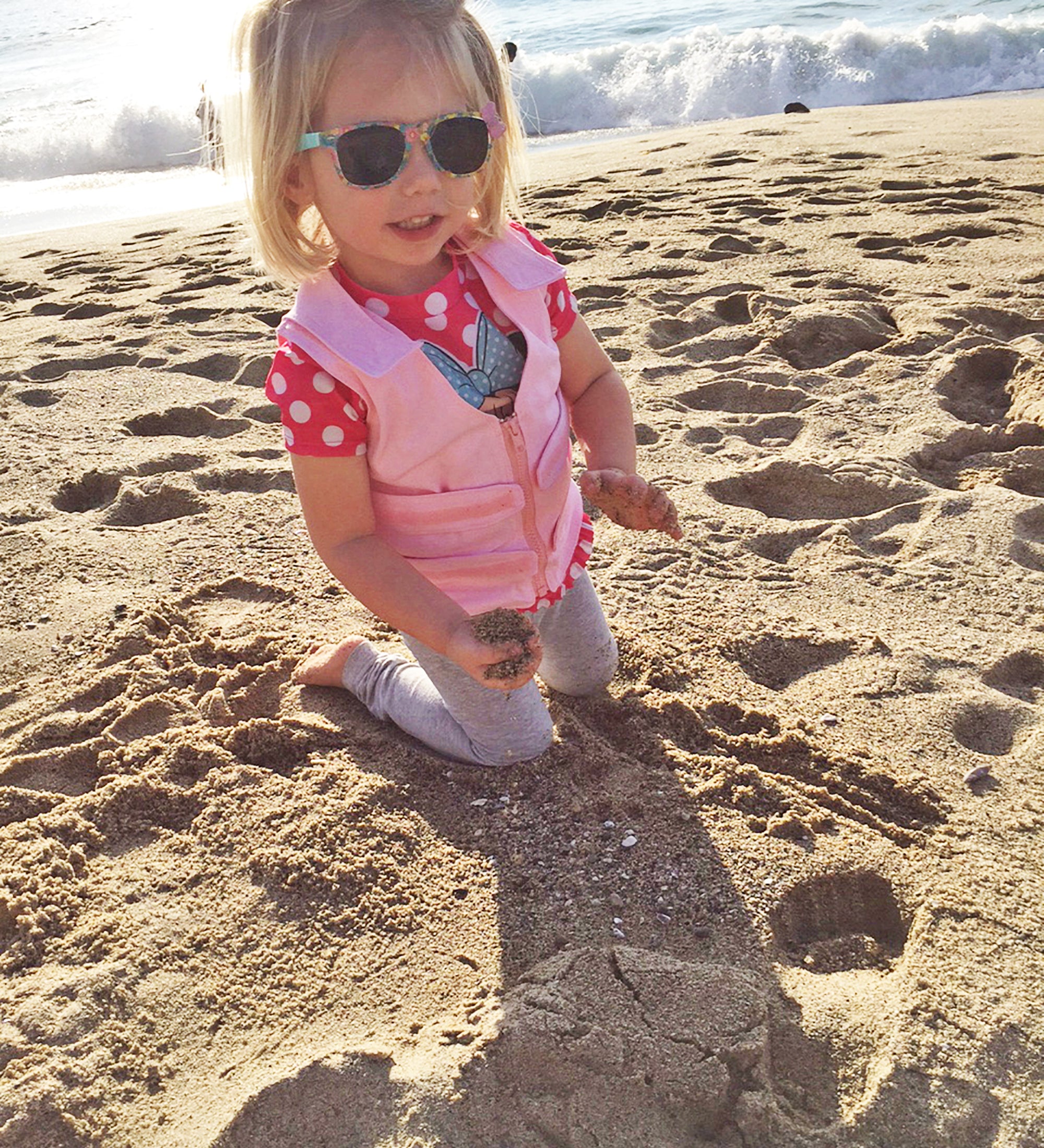 Child wearing sunglasses and a pink Cool Kids™ Toddler Cooling Kit with Vest, Neck Wrap, Extra Packs on a sandy beach with ocean waves in the background