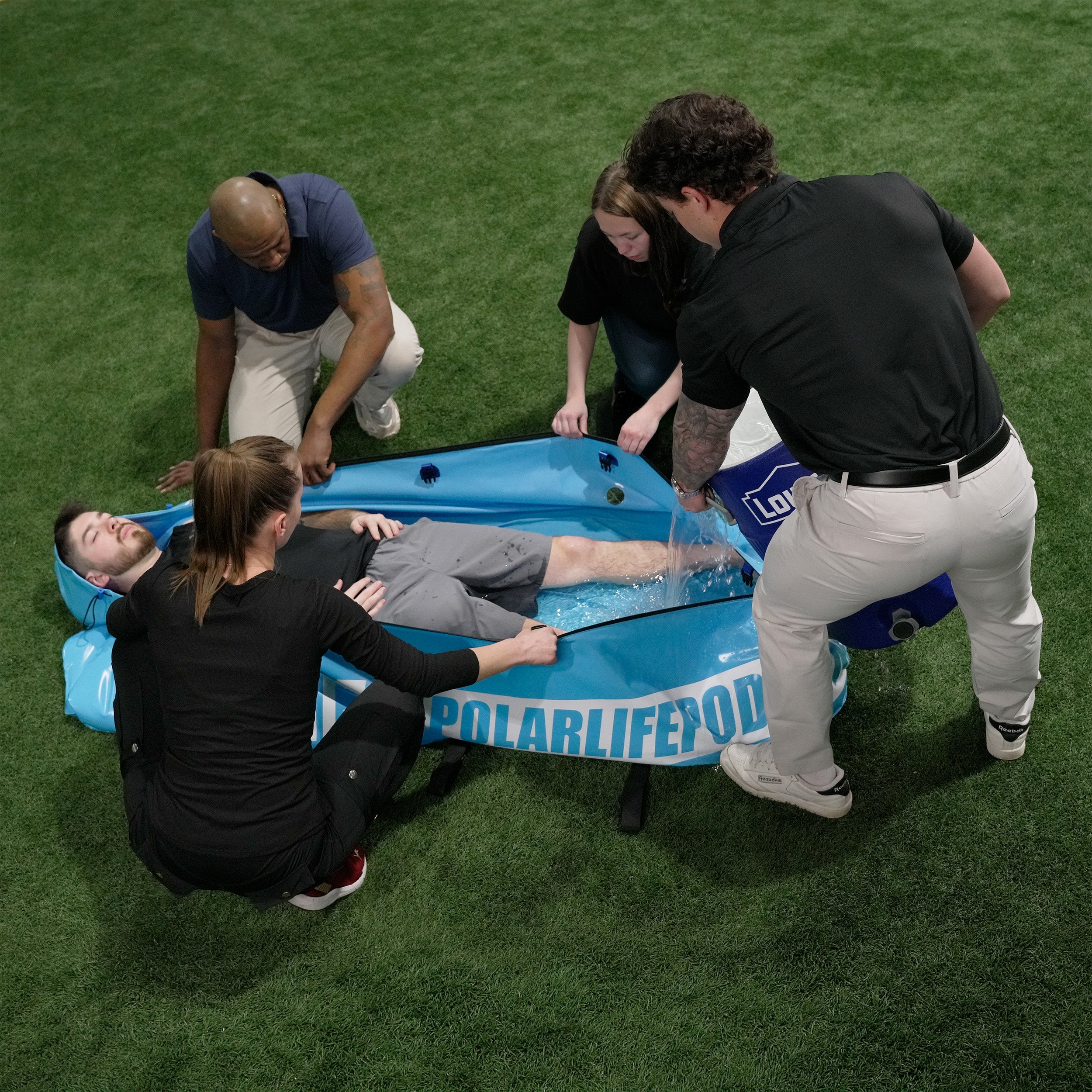 Person with heat illness laying in portable Polar Life Pod® with people holding it open and pouring in ice-water from 10 gallon container on a grassy field.