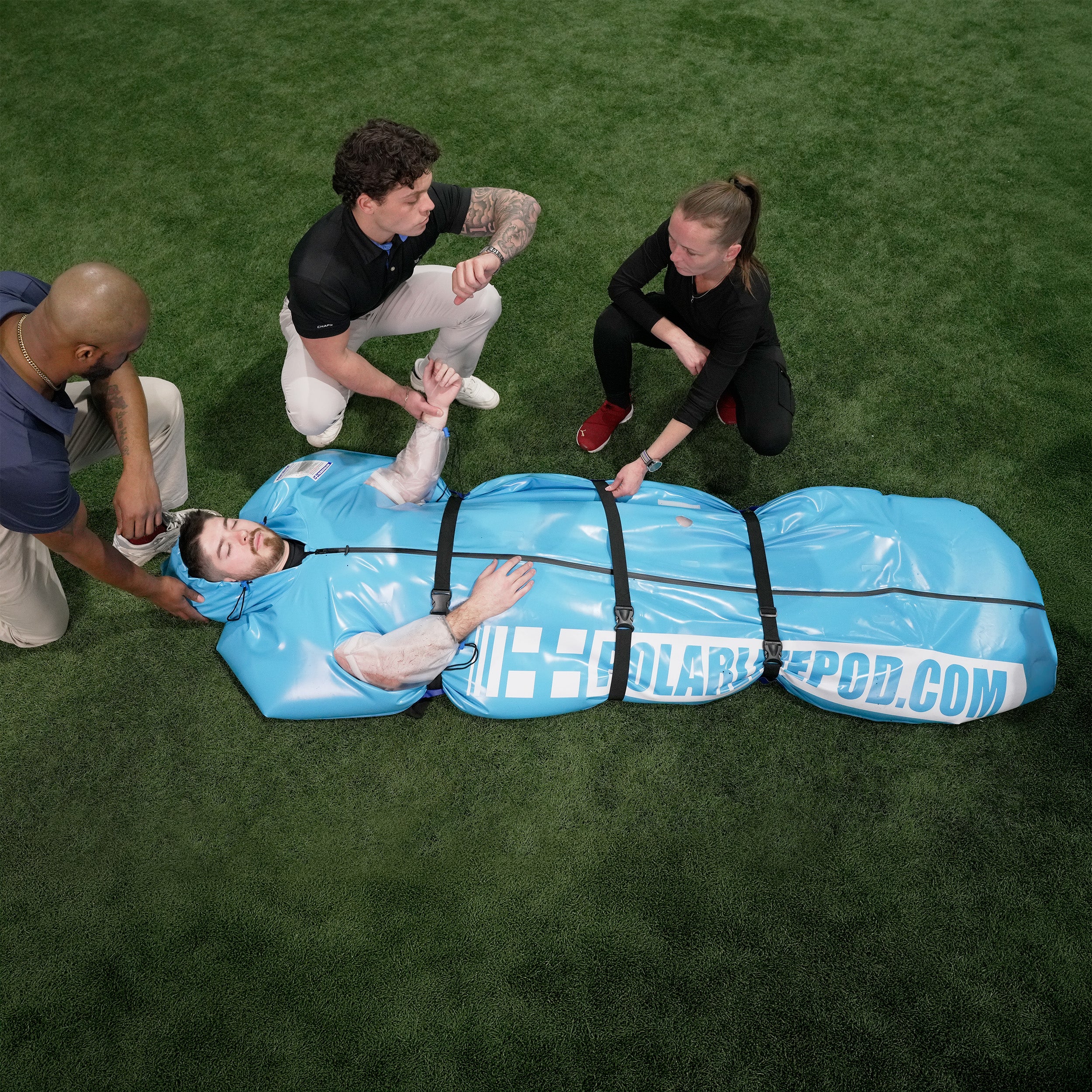 People assisting a person lying in the blue portable Polar Life Pod® on a grassy field.