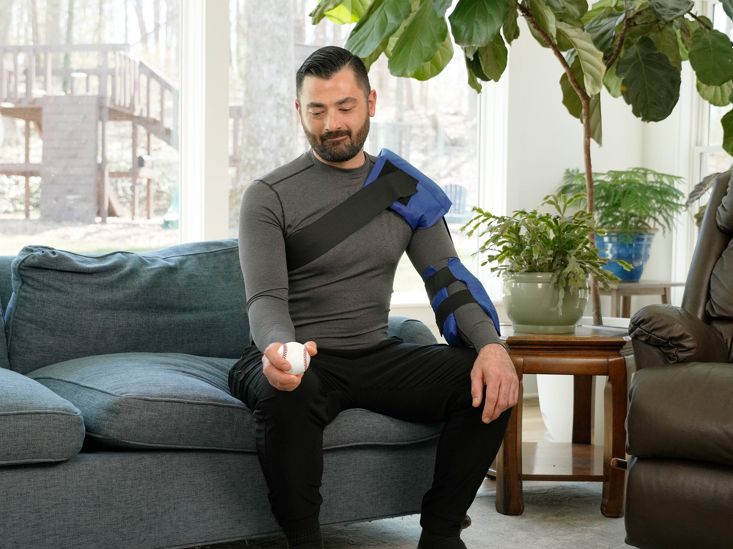 Man wearing a blue and black Soft Ice® Shoulder and Elbow Wrap in a living room, holding a baseball