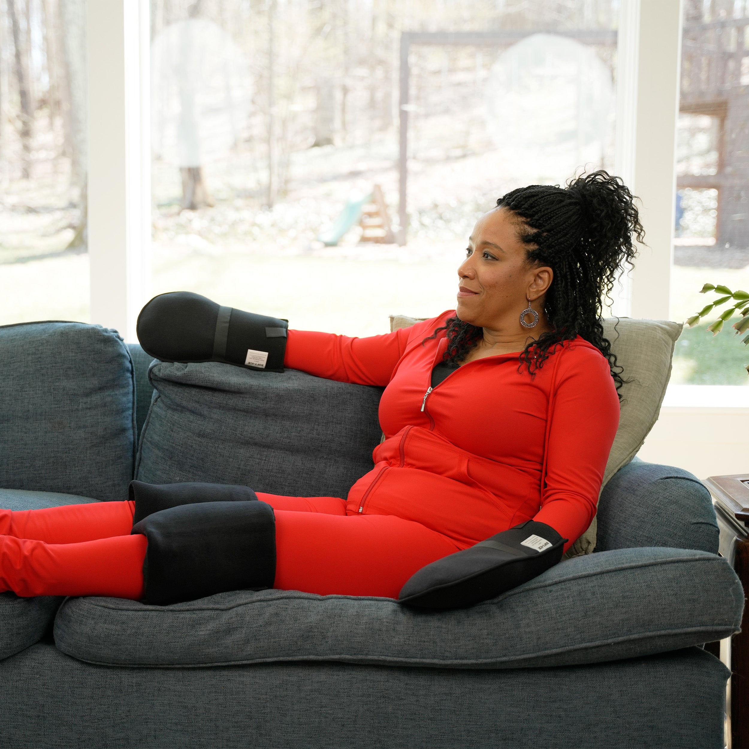 Woman in red outfit sitting on a gray couch in a living room wearing a Soft Ice® Compression Wrap Extremity Cooling Kit on her hands and knees.
