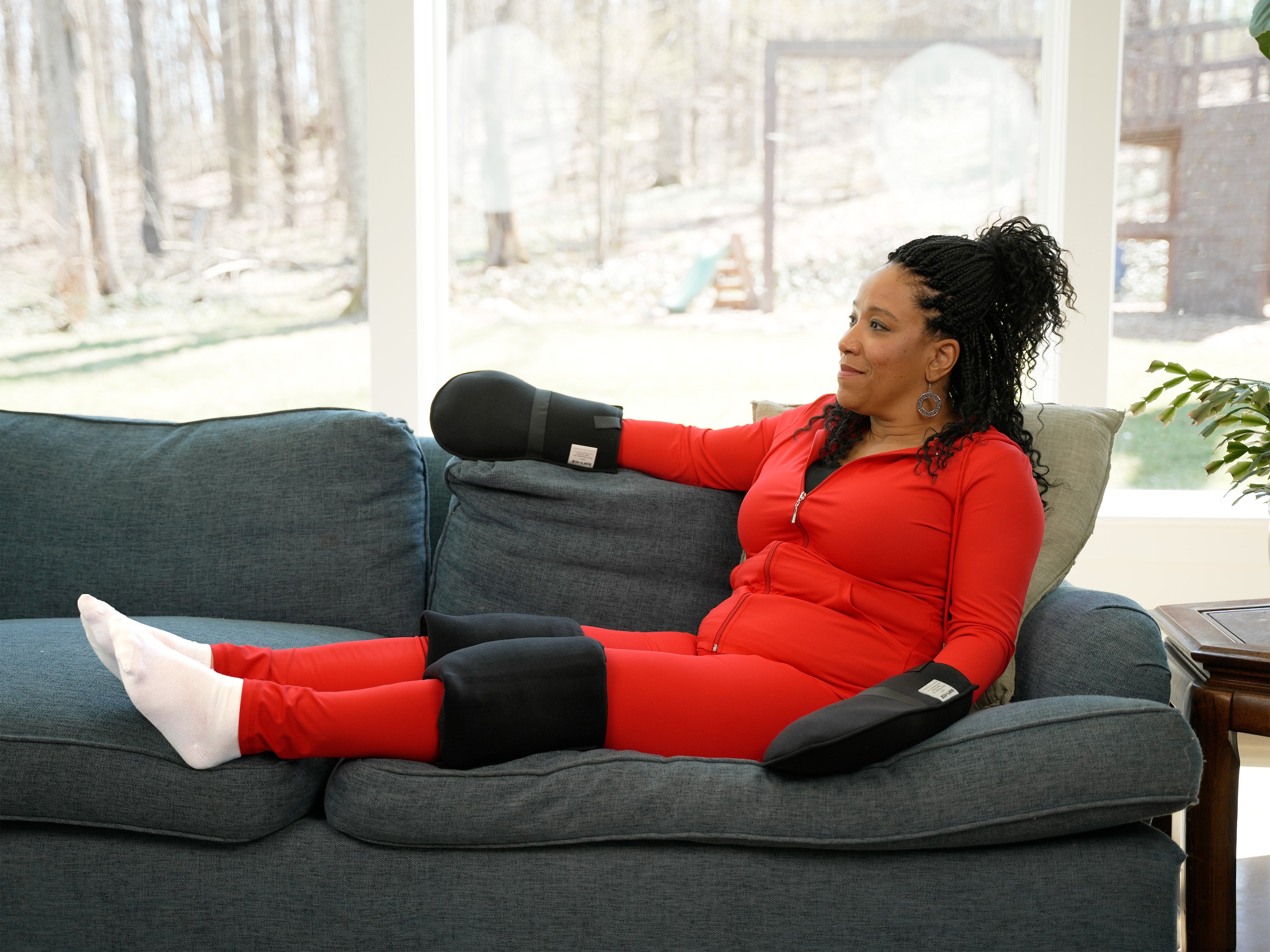 Woman in red outfit sitting on a gray couch in a living room wearing a Soft Ice® Compression Wrap Extremity Cooling Kit on her hands and knees.