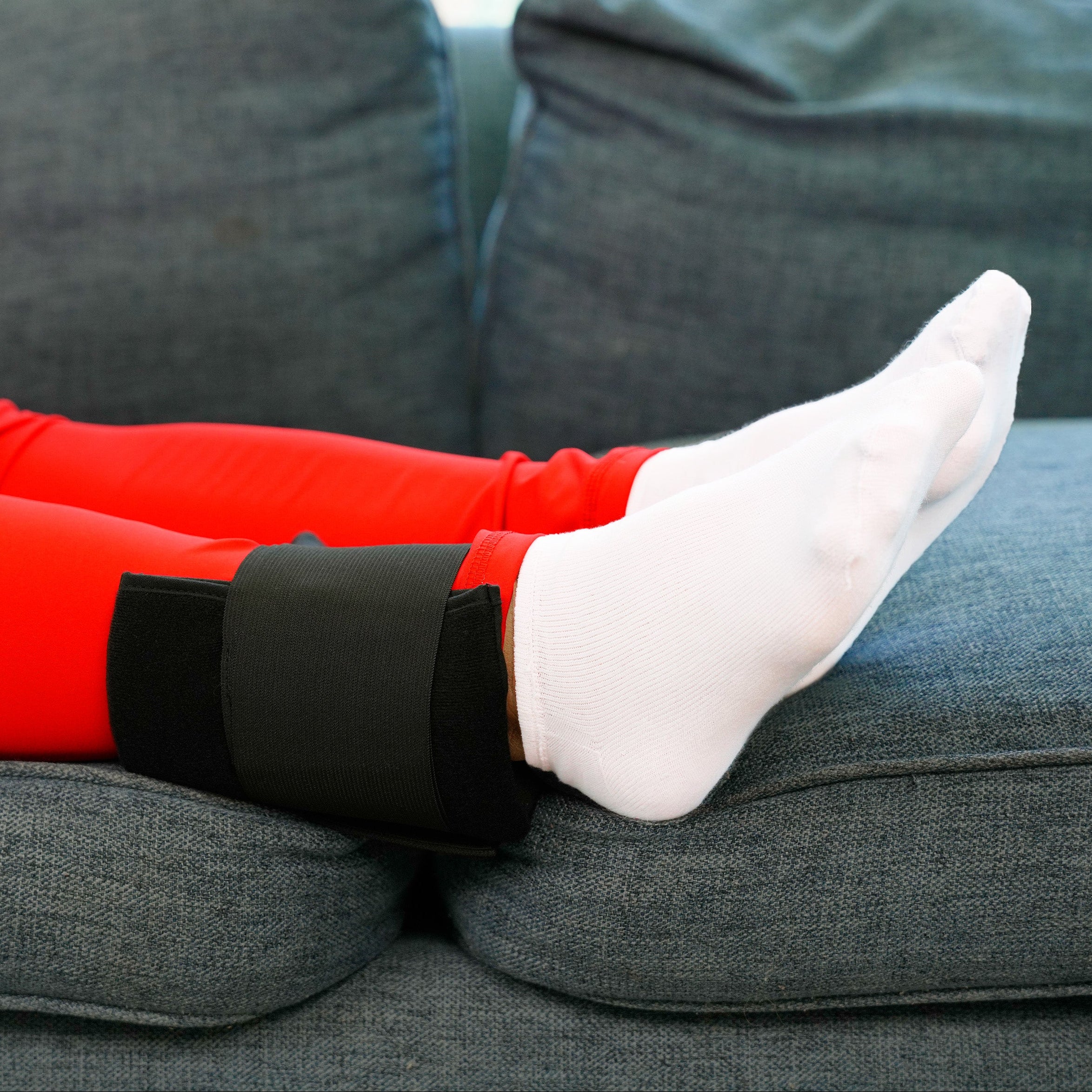 Person wearing red leggings with Soft Ice® Single Universal Compression Wrap on ankle sitting on a gray couch.