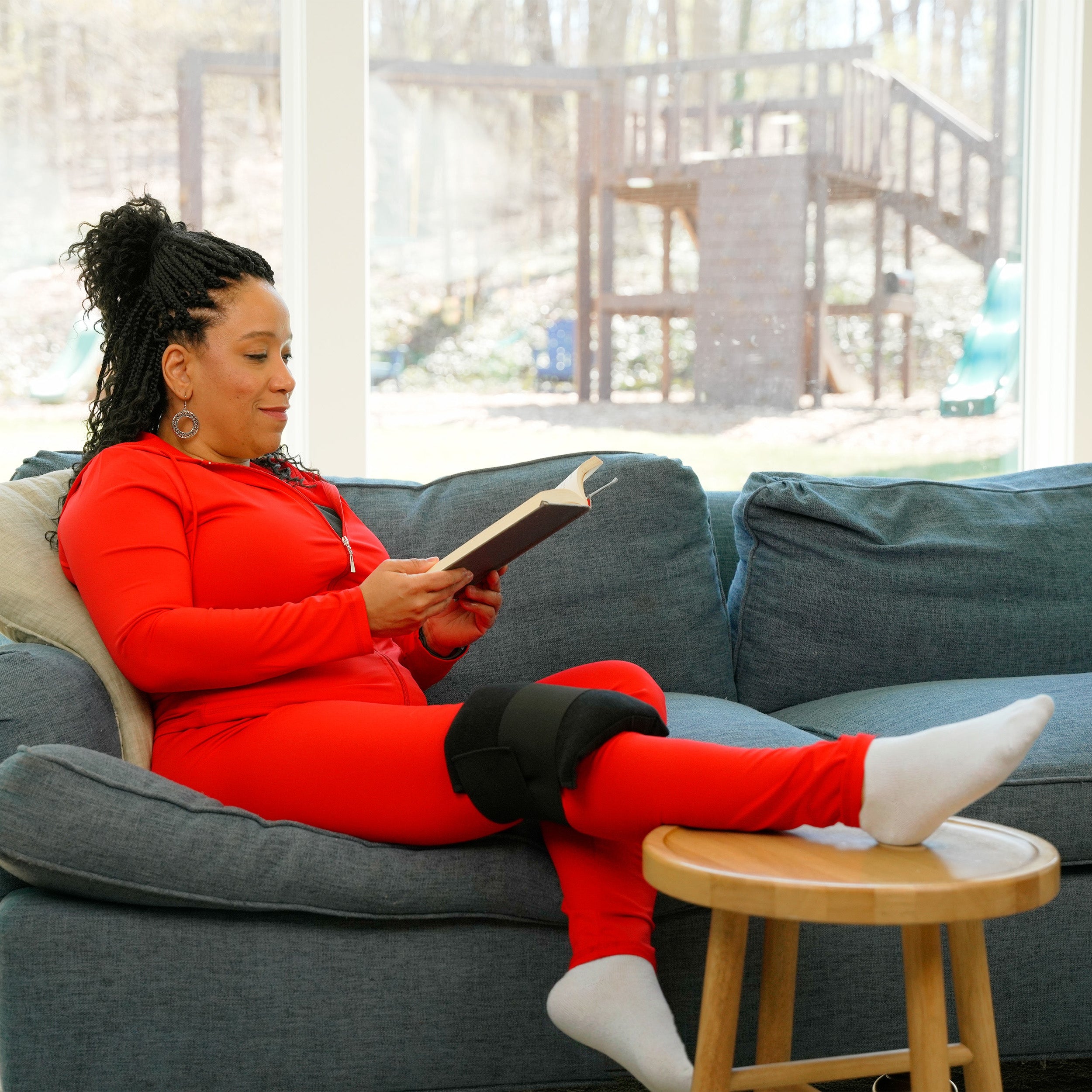 Woman in red outfit sitting on a gray couch reading a book in a bright room wearing a Soft Ice® Single Universal Compression Wrap on her knee.