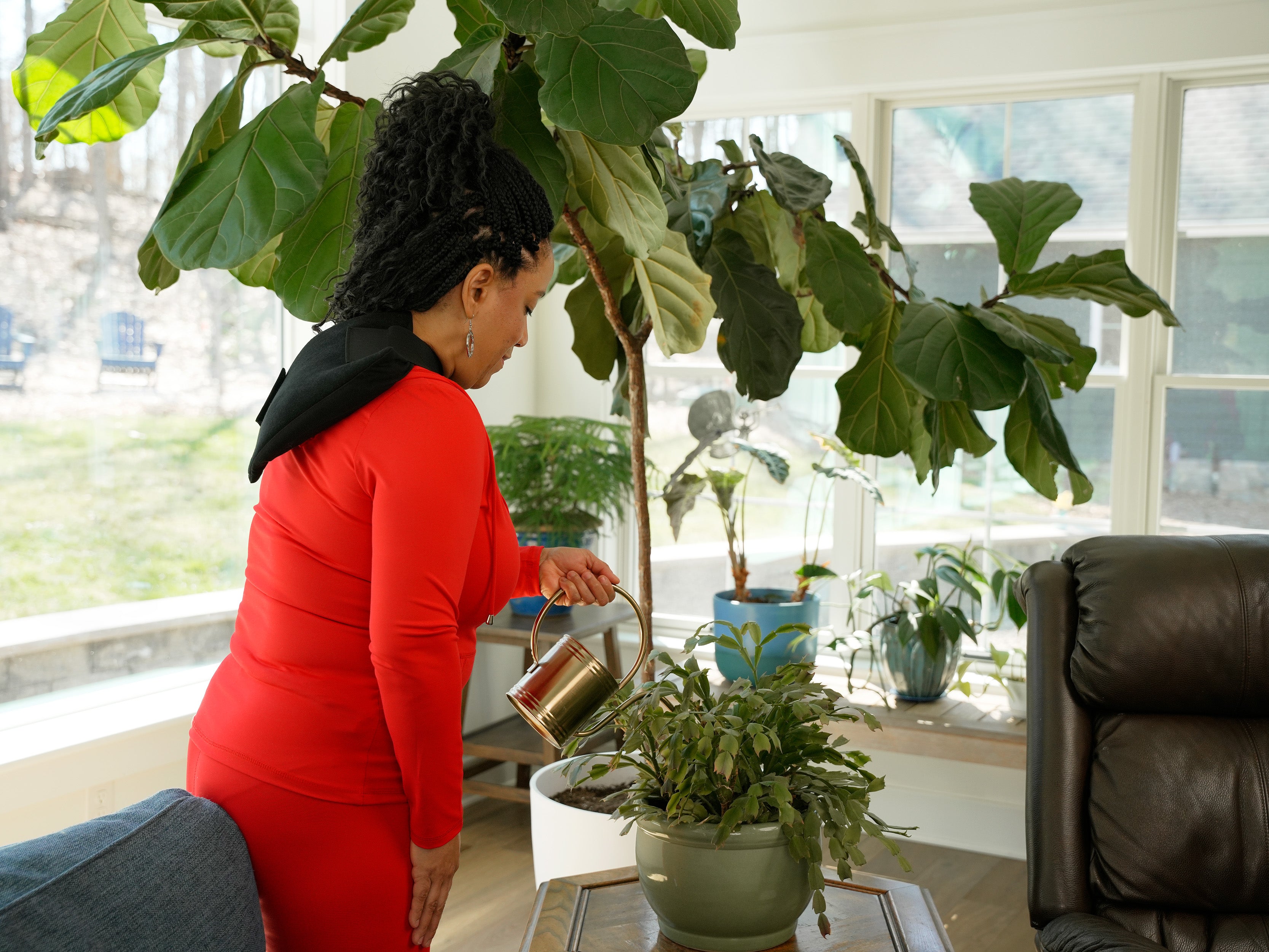 A woman in a red long-sleeve outfit with a black deluxe single universal soft ice® wrap on her shoulder, pouring water from a metallic watering can into a potted green plant on a table inside a bright room with large windows and indoor plants.