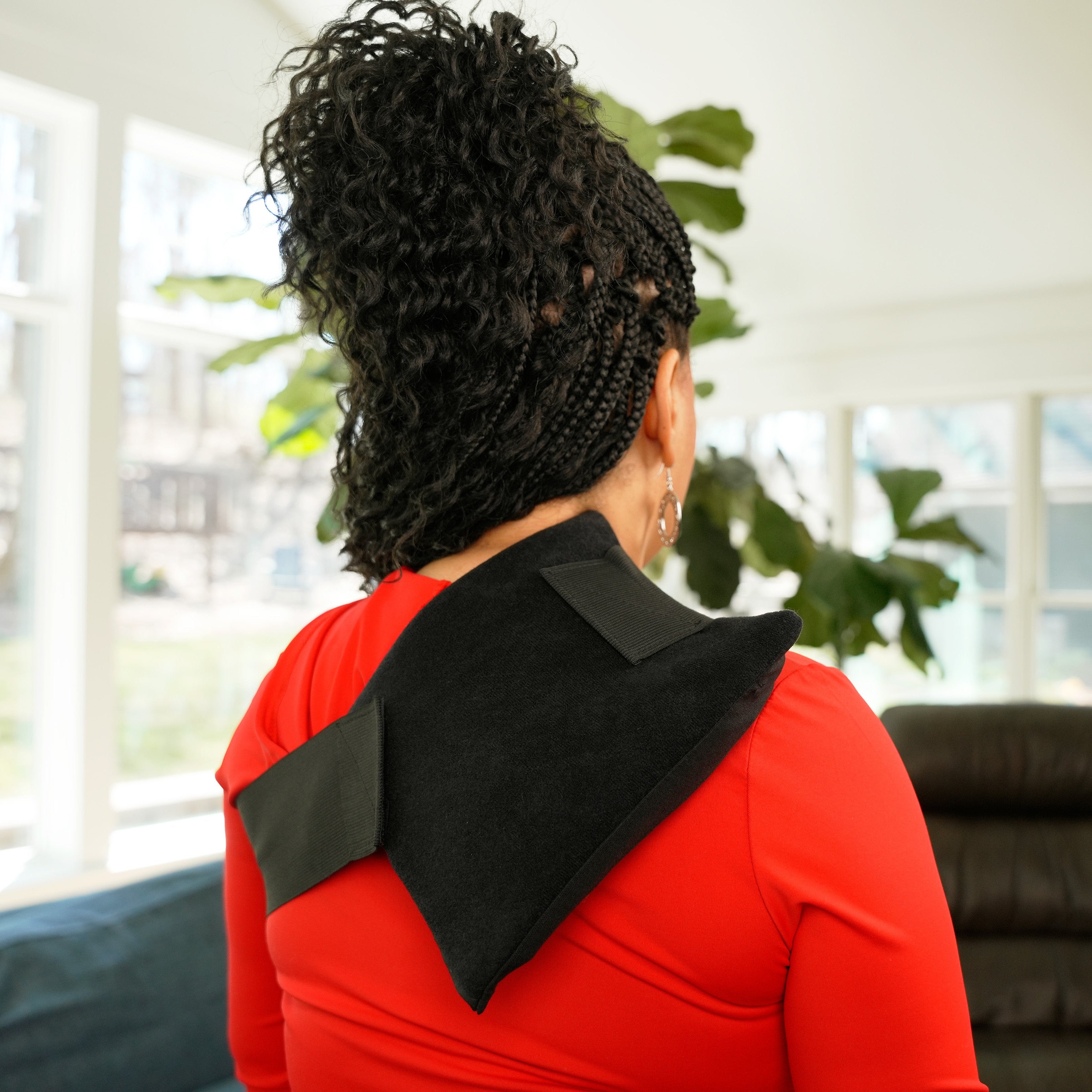 A woman with curly black hair is shown from behind wearing a black deluxe single universal soft ice wrap over a bright red top in a modern living room setting.