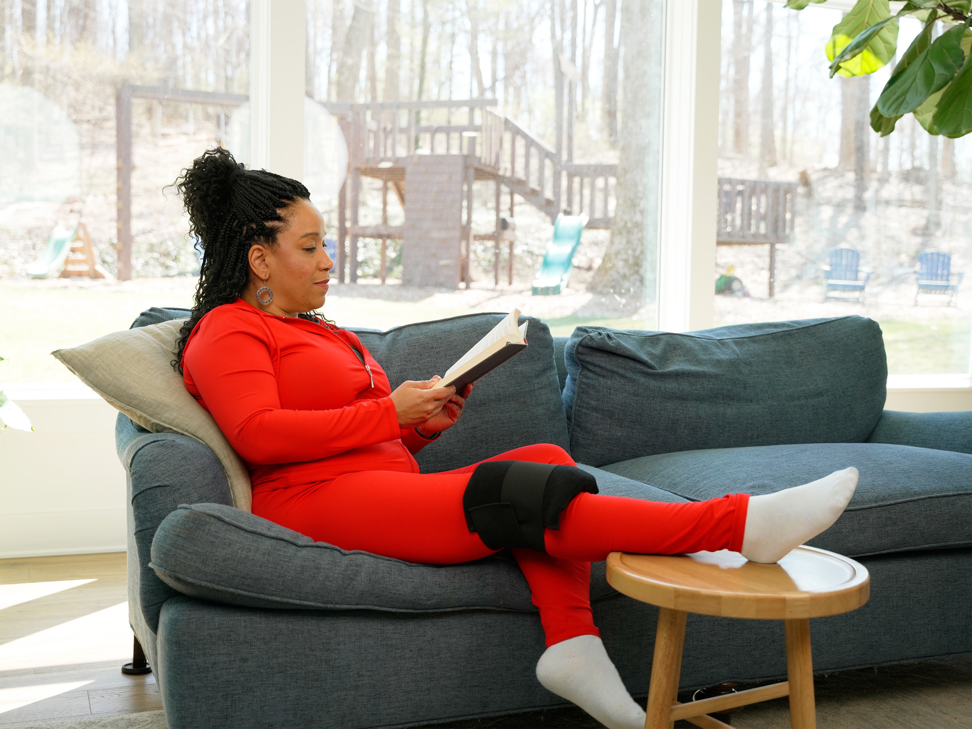 A woman in a red matching activewear set sits on a blue-gray couch, reading a book with one leg resting on a small wooden stool. She has a black deluxe single universal soft ice® wrap on her right leg, and the background shows a large window looking out onto a backyard play area.