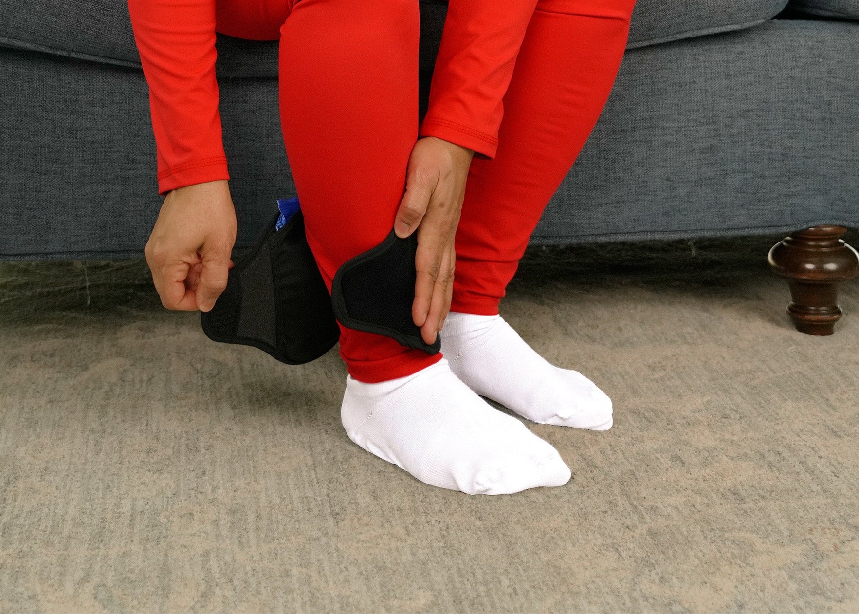 Woman in red outfit sitting on a couch applying black Soft Ice® Ankle Wrap to her ankle.