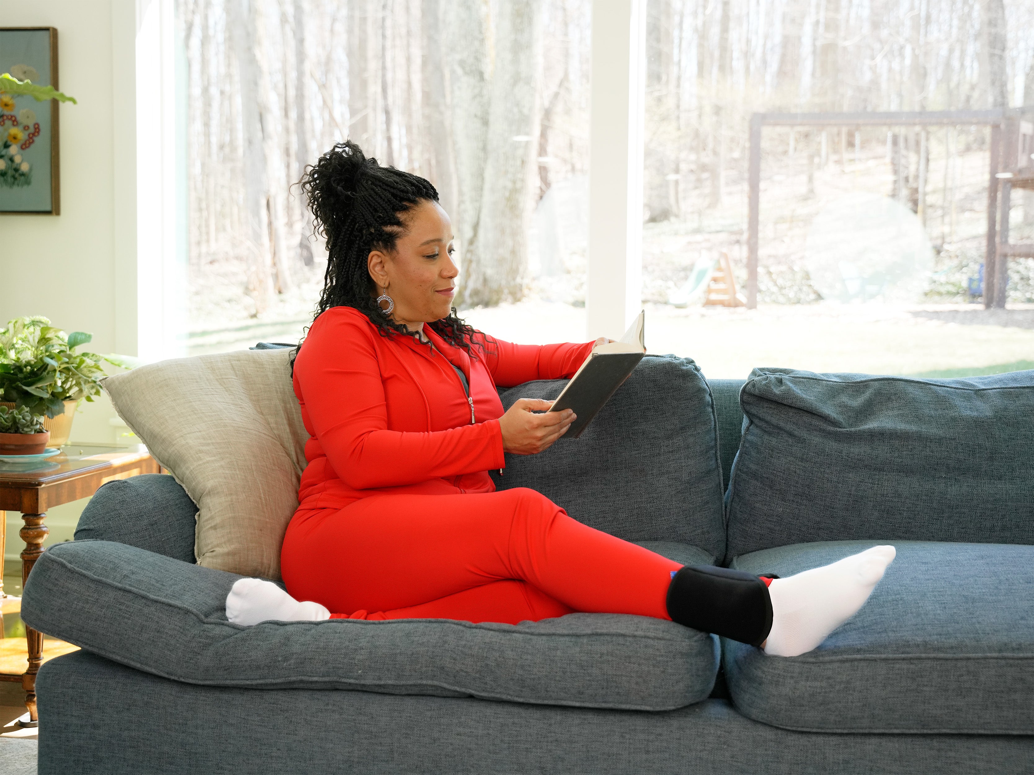 Woman in red outfit sitting on a gray couch reading a book in a living room wearing a black Soft Ice® Ankle Wrap.