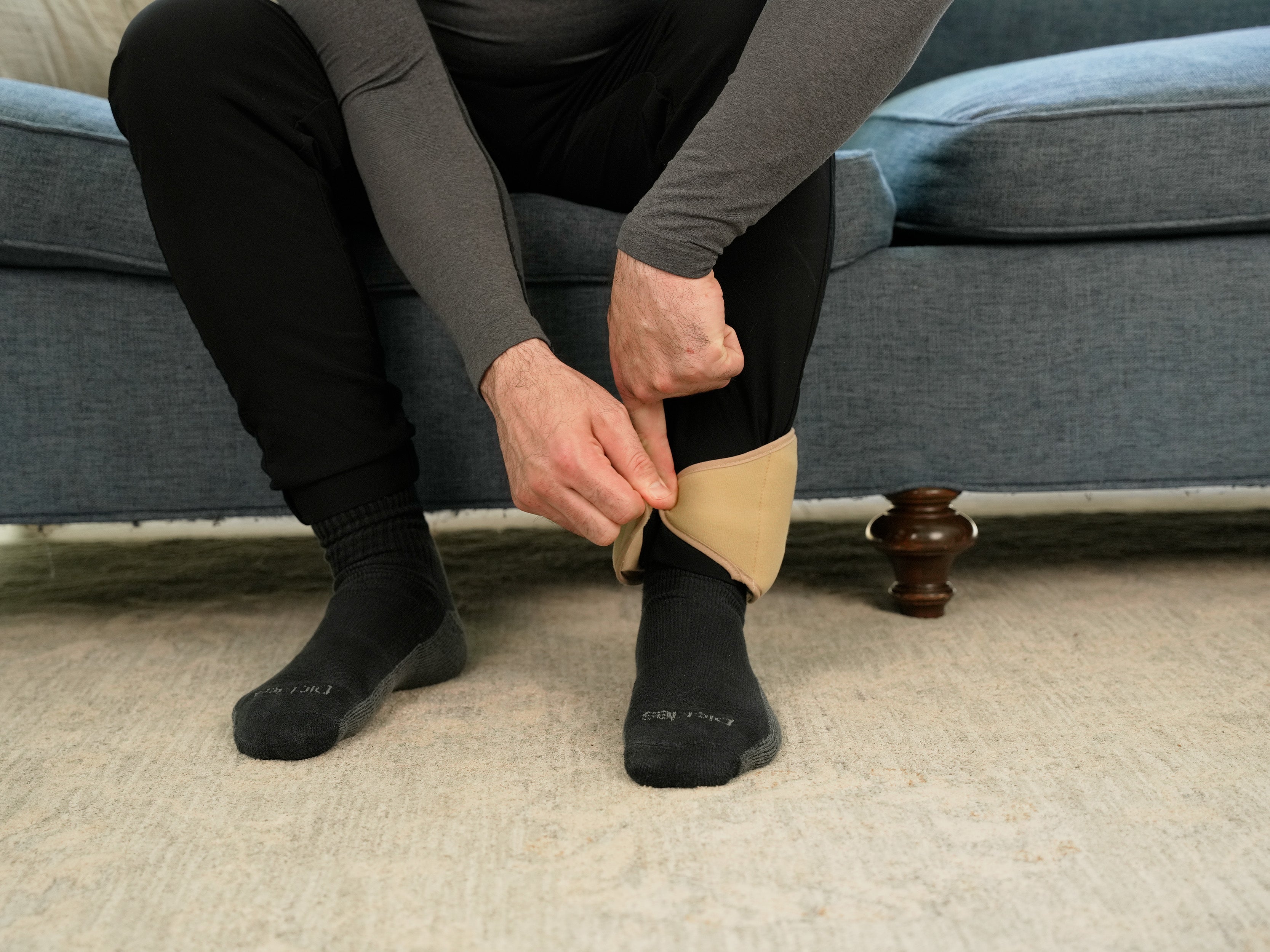 Person sitting on a couch wearing black socks applying a Khaki Soft Ice® Ankle Wrap.