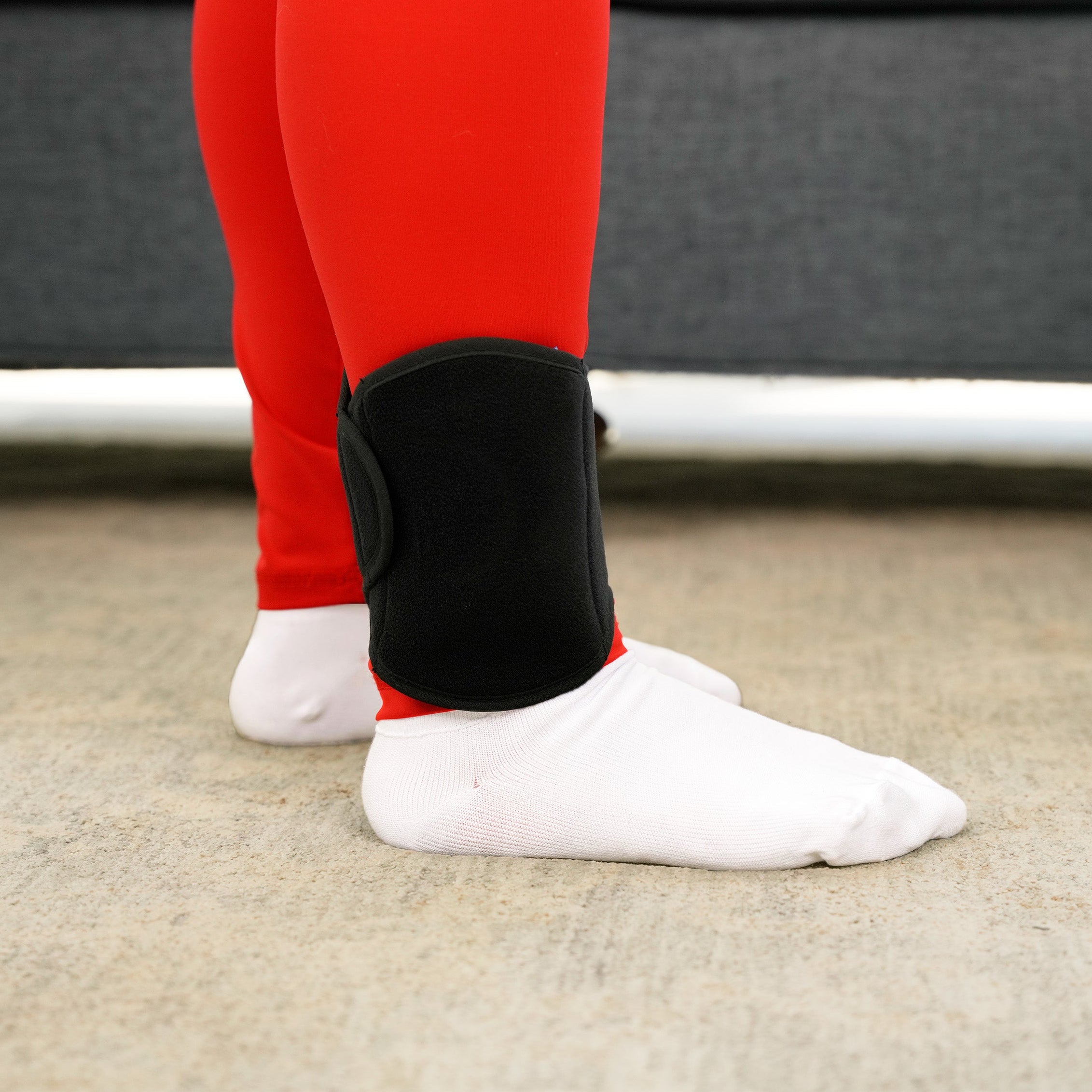 Person wearing red leggings, white socks, and a black Soft Ice® Ankle Wrap on a carpeted floor.