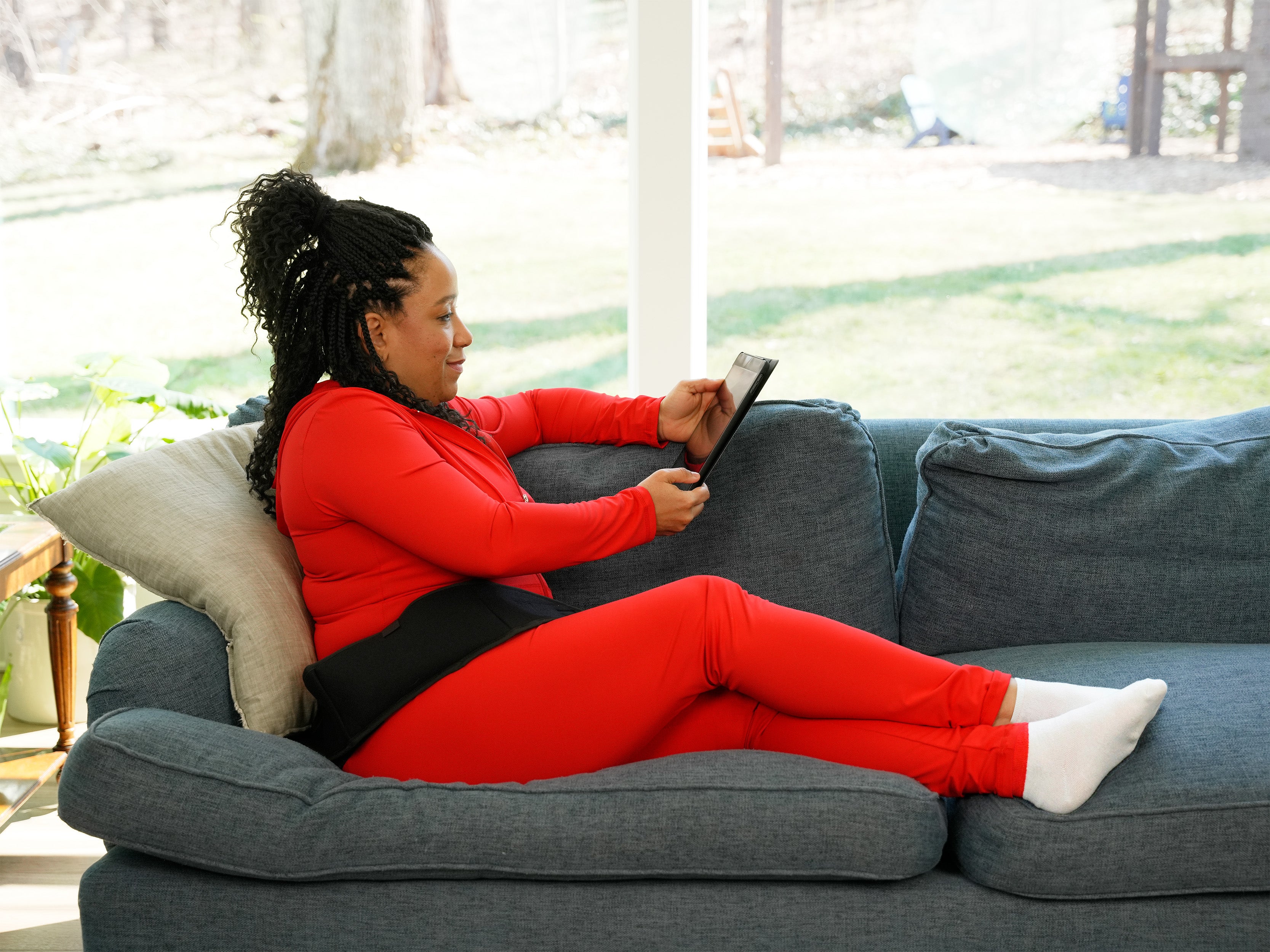 Woman in red outfit sitting on a gray couch using a tablet wearing a Soft Ice® Standard Universal Compression Wrap on her hip.