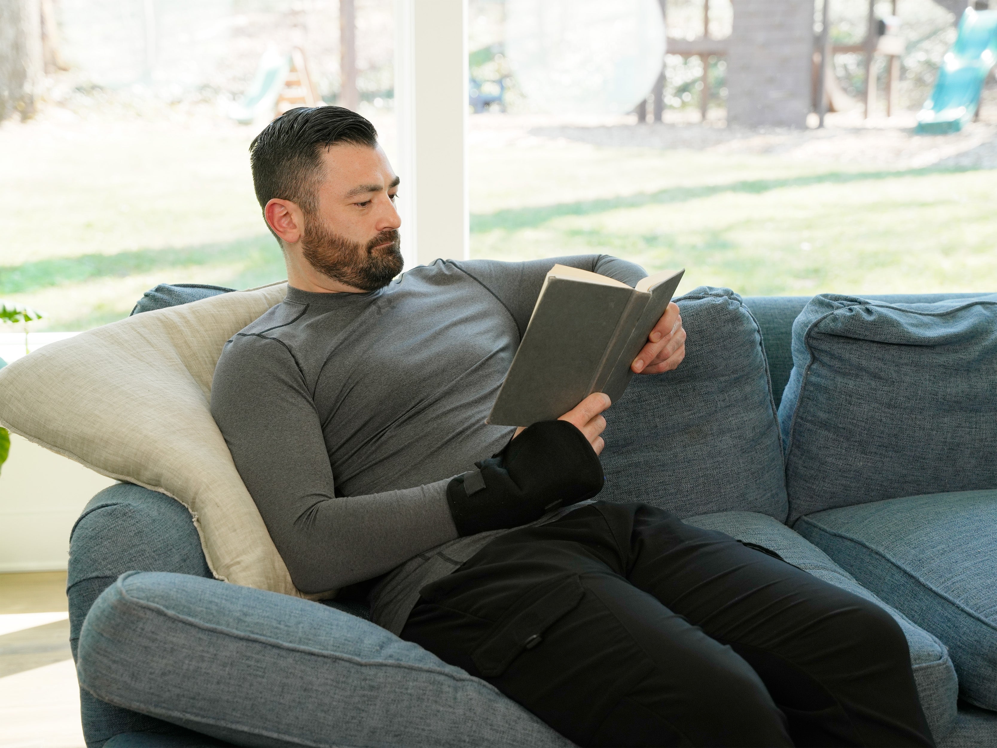 Man reading a book on a blue couch in a bright living room wearing a Soft Ice® Wrist Wrap.