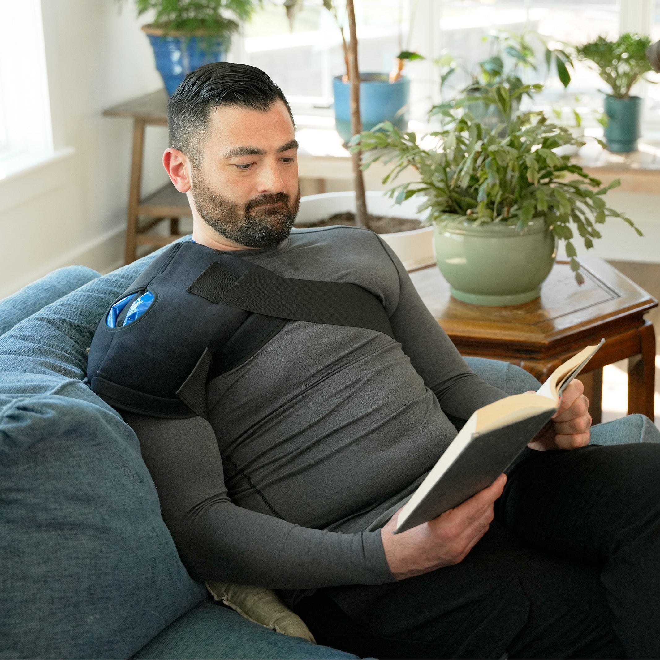 Man reading a book on a blue couch in a living room with plants and a leather chair wearing a Soft Ice® Shoulder Wrap.
