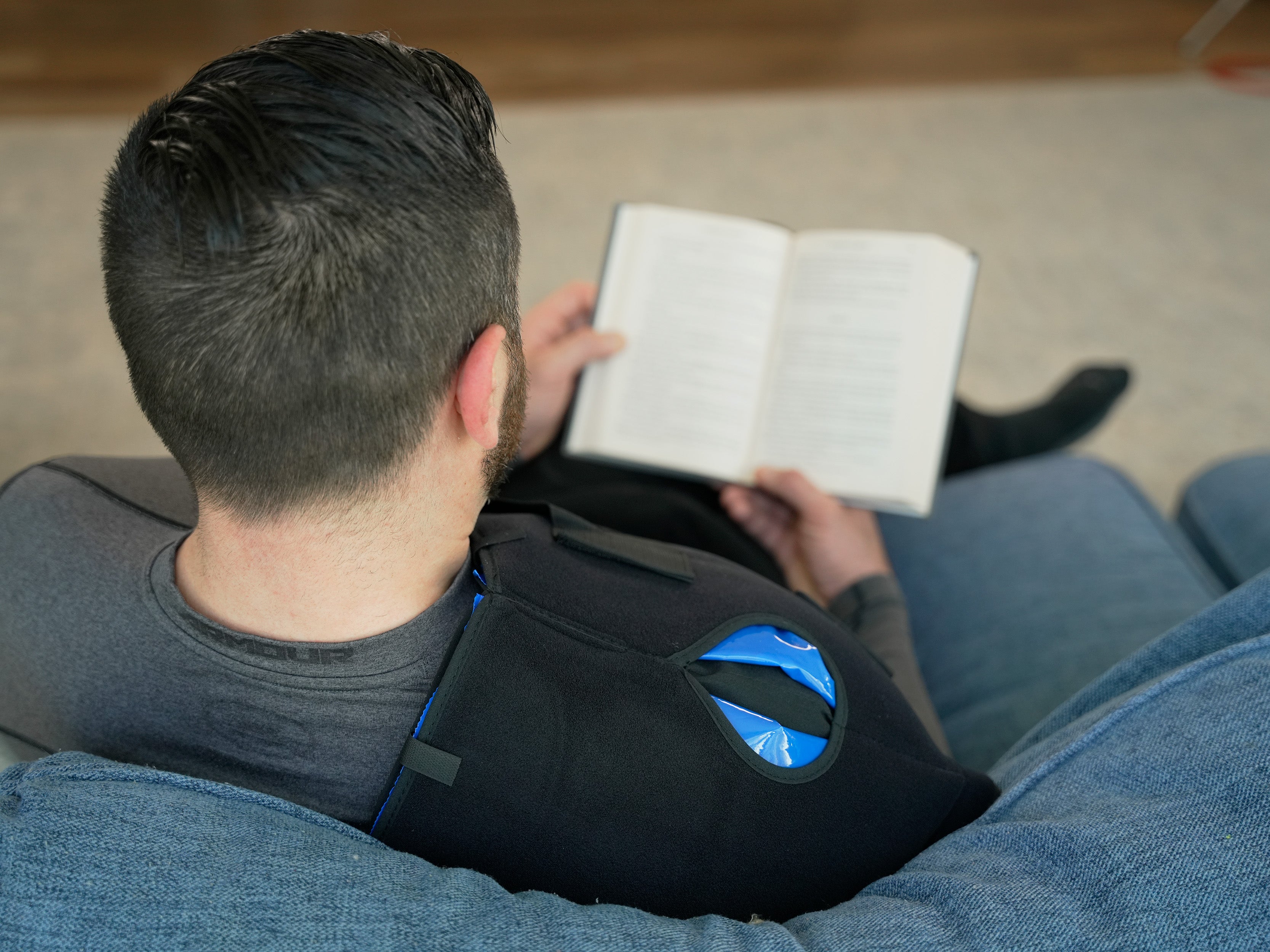 Person sitting on a couch reading a book with a Soft Ice® Shoulder Wrap on