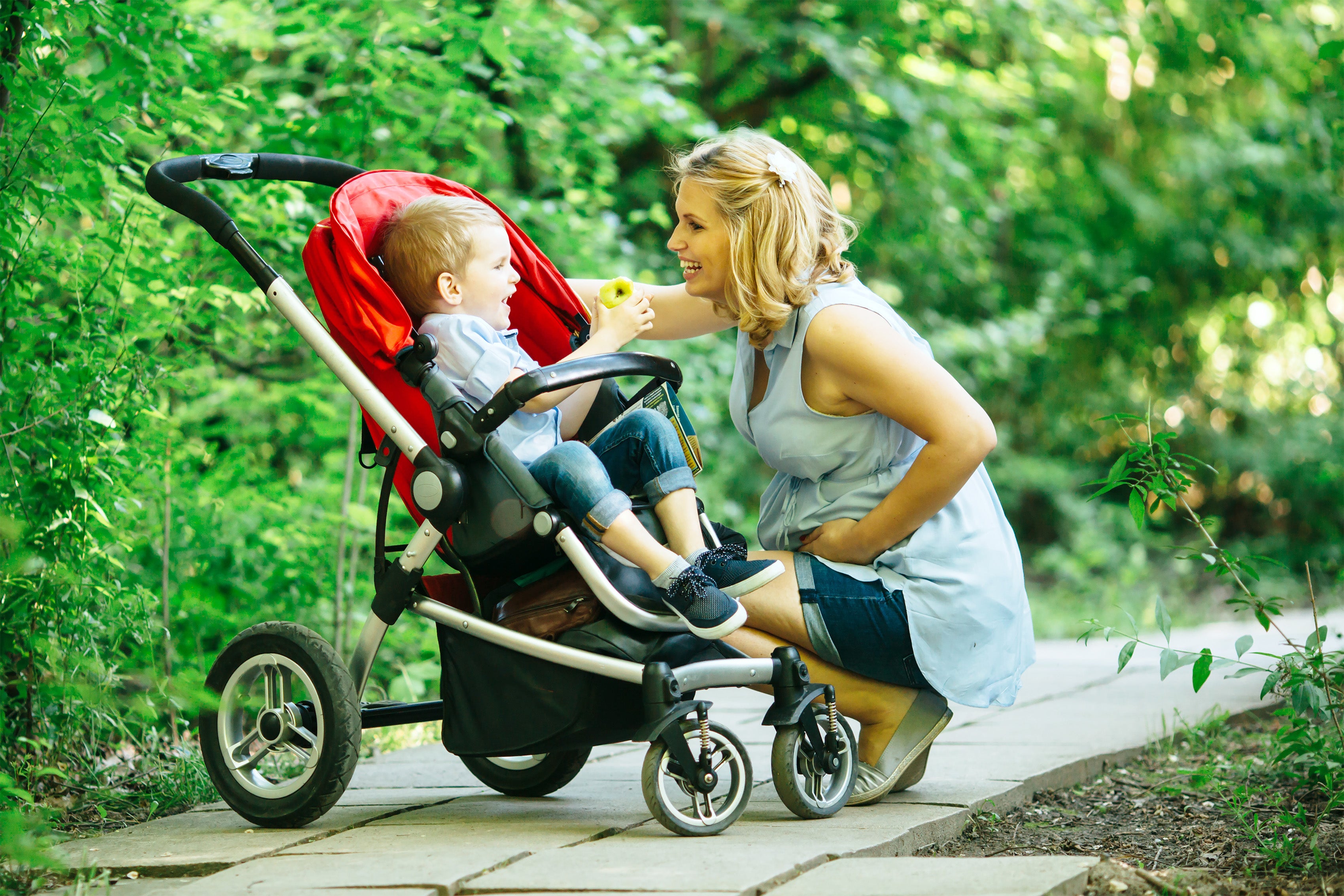 Woman laughing with her child in his stroller outside on a path while he's eating an apple. 