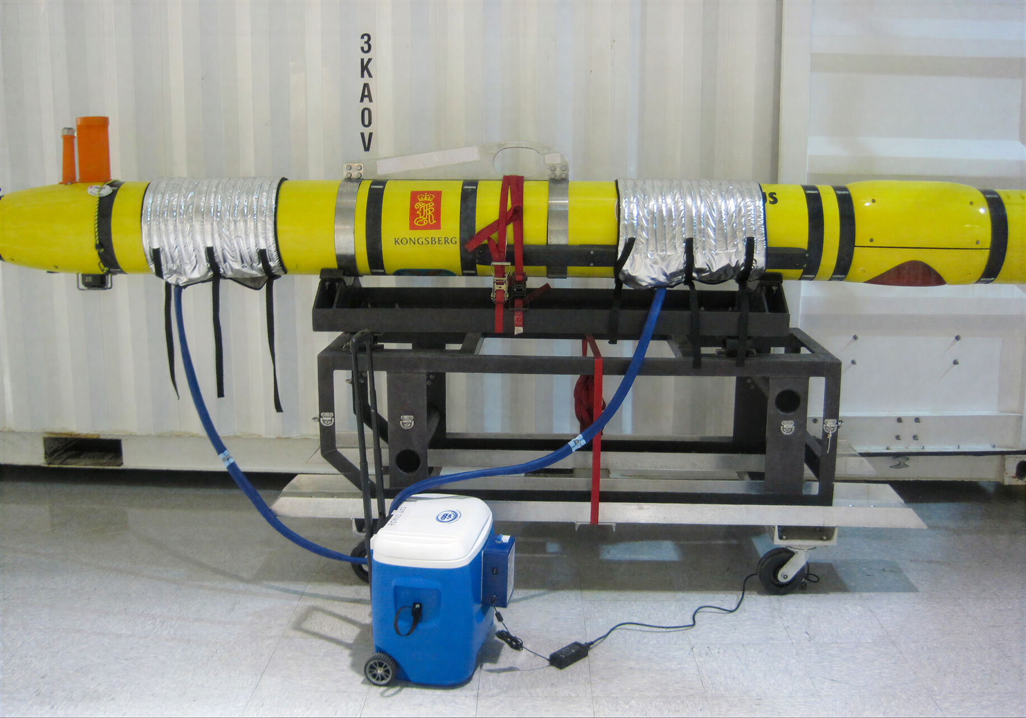 A yellow underwater torpedo or missile mounted on a black metal support frame inside a white container or lab space. Blue hoses connect the submarine to a blue and white cooling unit on the floor.