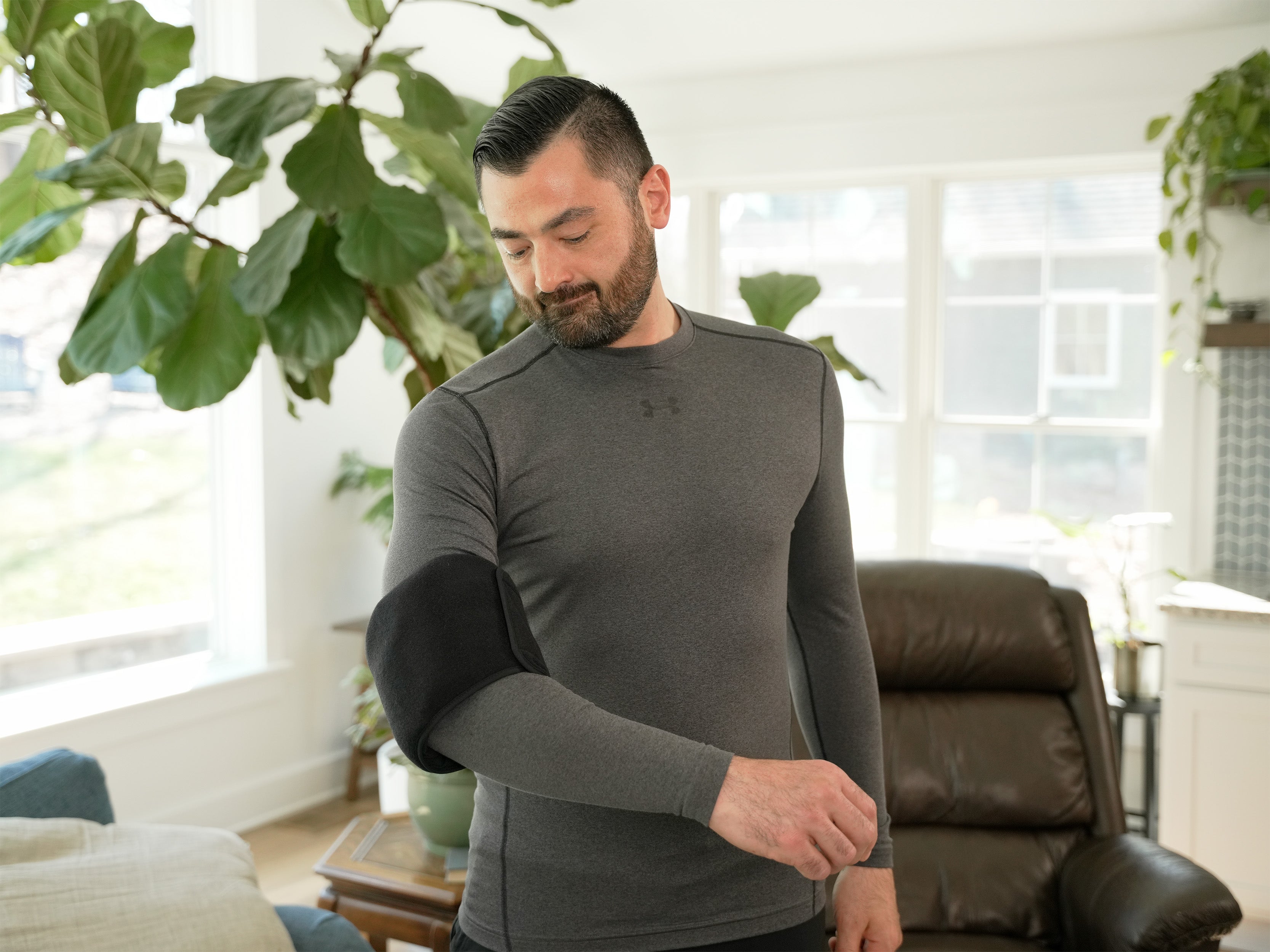 Man wearing a gray long-sleeve shirt in a living room setting with plants and furniture wearing a Soft Ice® Double Universal Compression Wrap on his arm.