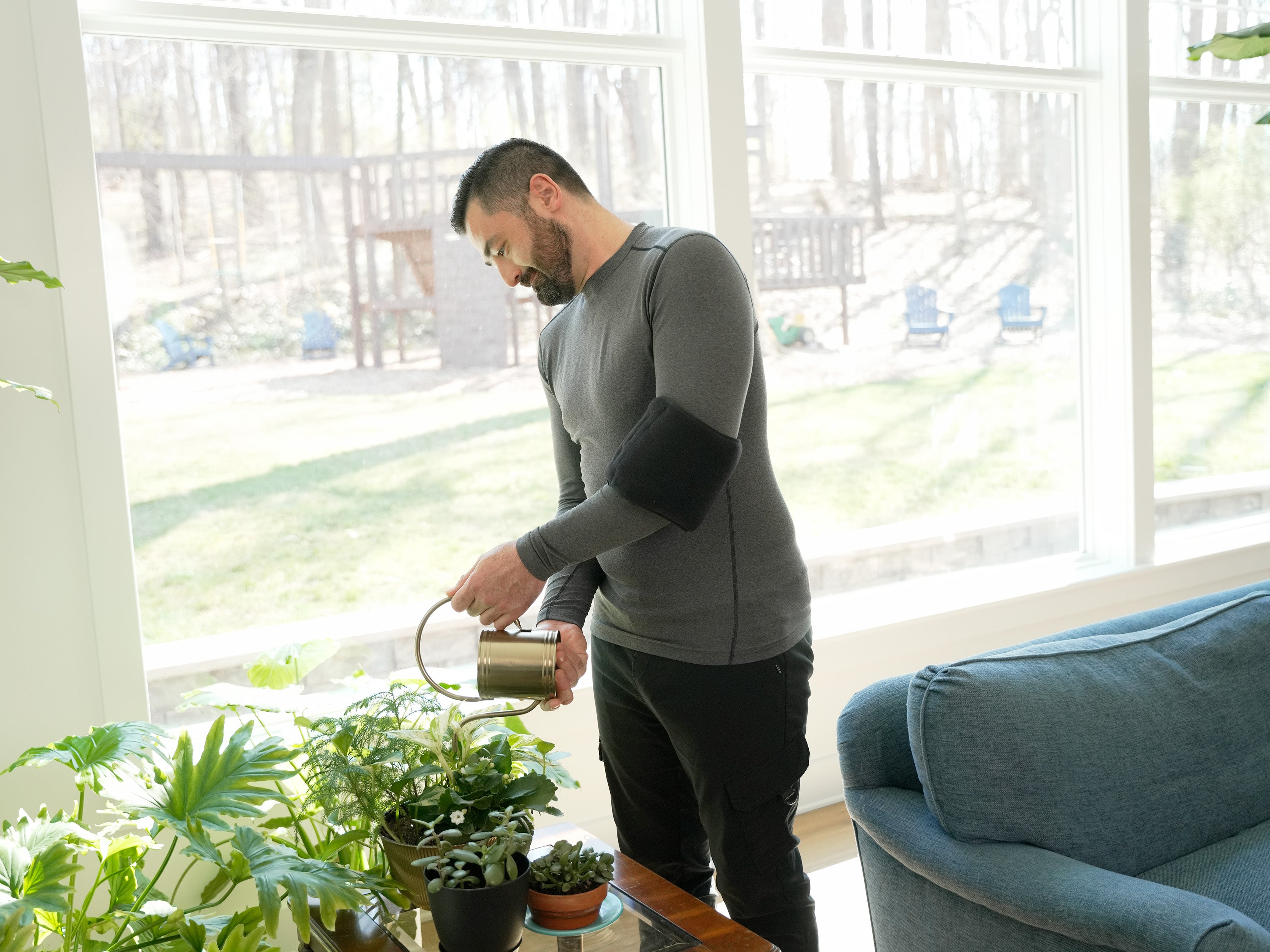 Man watering plants in a room with large windows wearing a black Soft Ice® Double Universal Compression Wrap on his arm