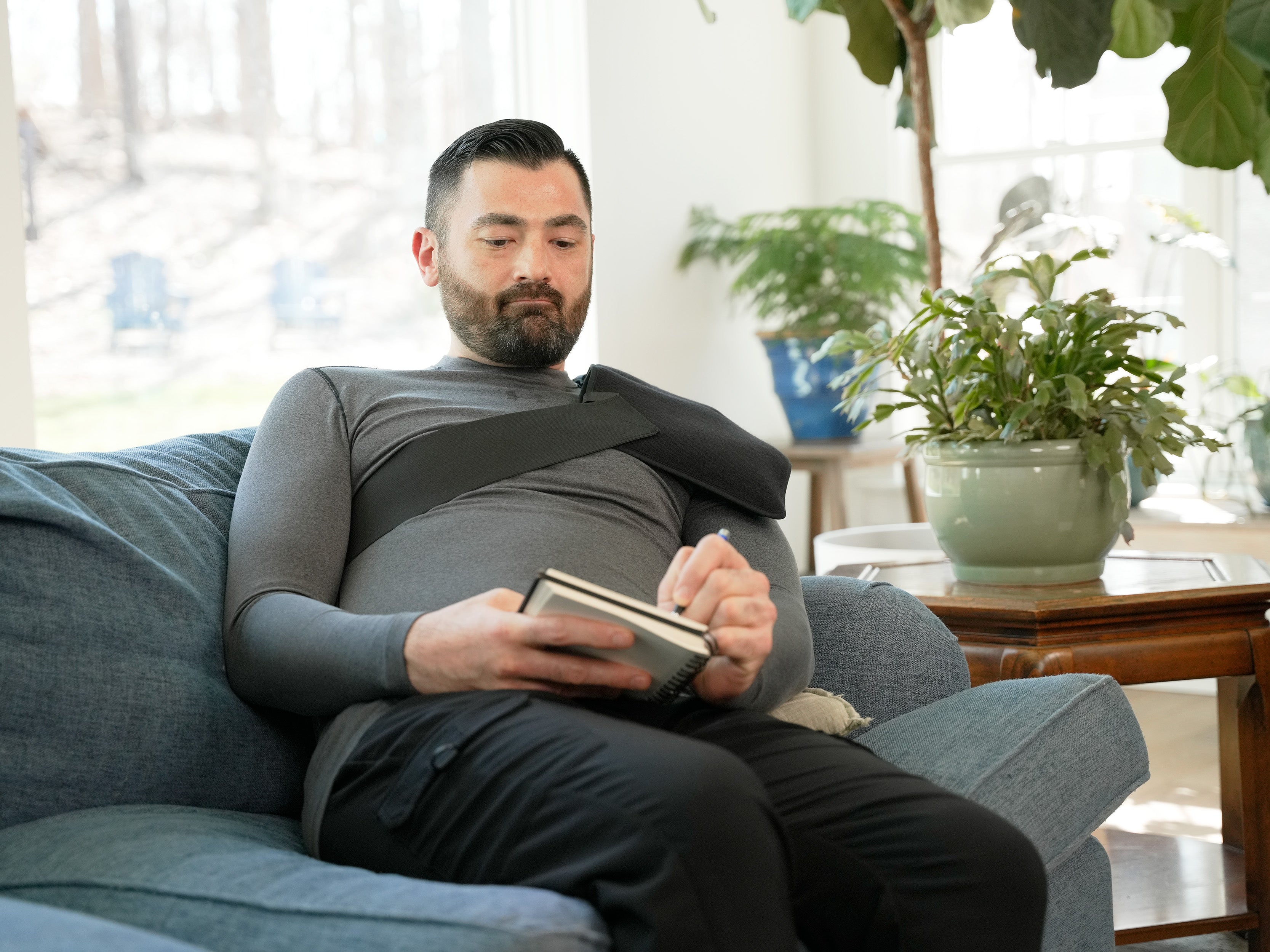 Man sitting on a couch with a book, wearing a Soft Ice® Triple Universal Compression Wrap on his shoulder, in a home setting with plants.