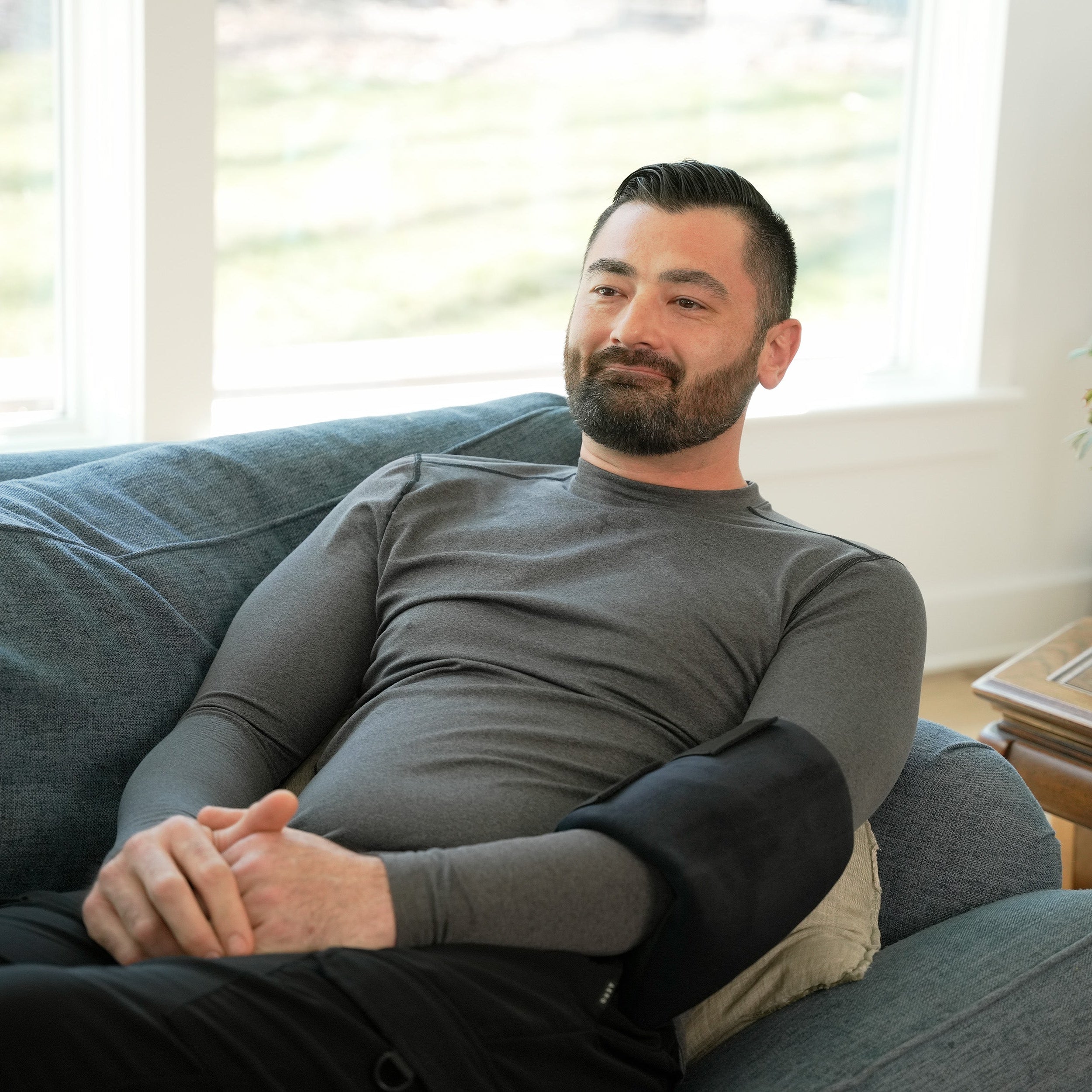 Man sitting on a couch in a bright living room with plants and a window in the background wearing a Soft Ice® Large Universal Joint Compression Wrap on his elbow.