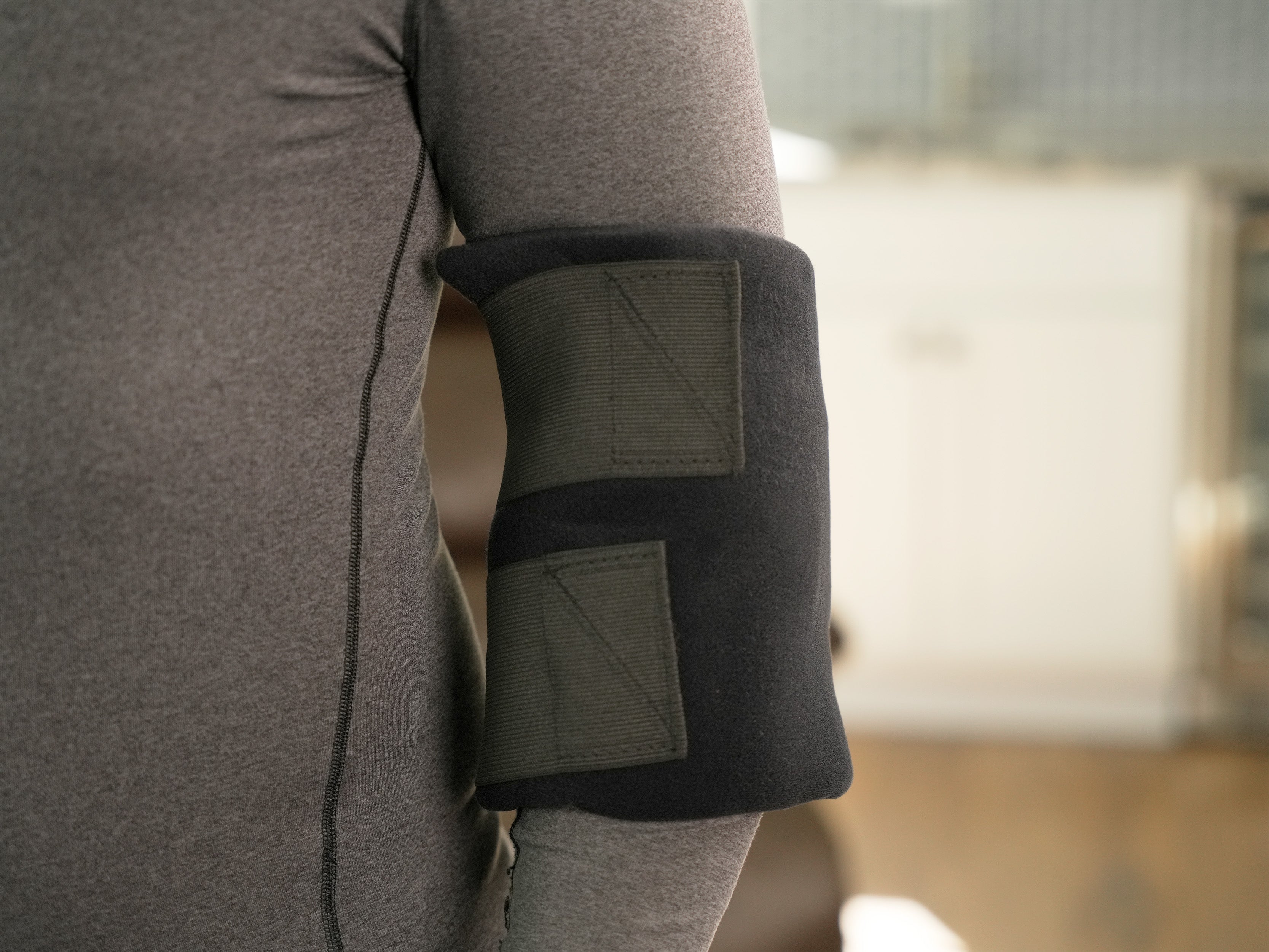 Man wearing a Soft Ice® Large Universal Joint Compression Wrap on his elbow in a blurred living room setting.