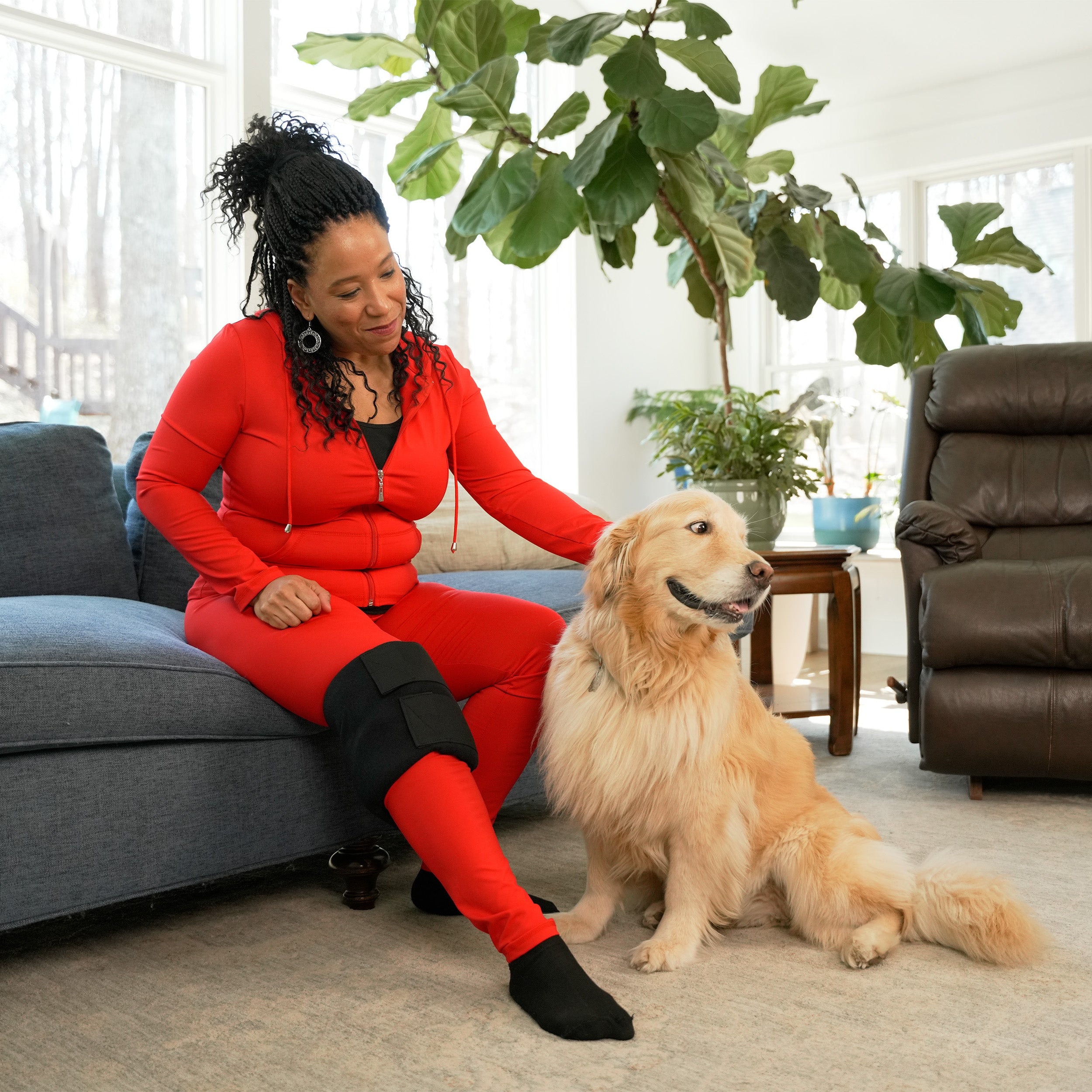 Woman in red outfit sitting on a couch wearing a Soft Ice® Medium Universal Joint Compression Wrap on her knee with a golden retriever in a living room.