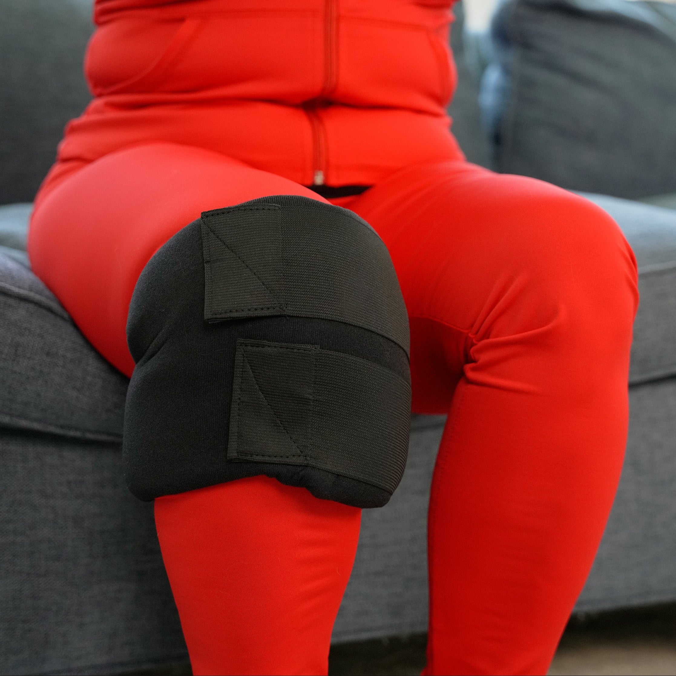 Person wearing a red outfit with a black Soft Ice® Medium Universal Joint Compression Wrap on her knee sitting on a gray couch.
