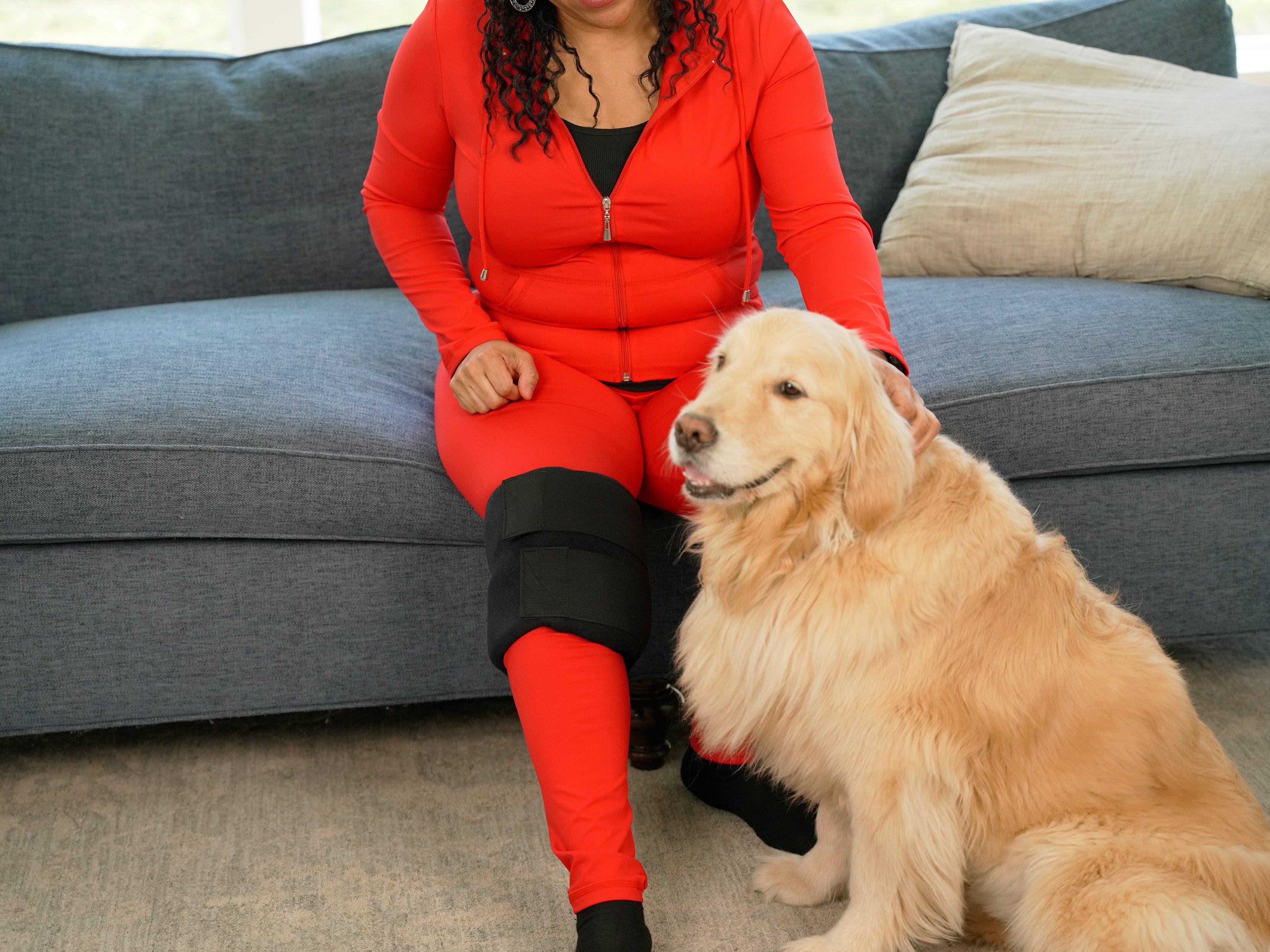 Person in red outfit sitting on a gray couch wearing a Soft Ice® Medium Universal Joint Compression Wrap on her knee with a golden retriever dog.