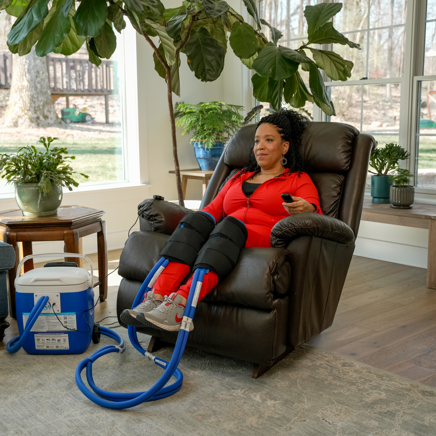 A woman reclining in a brown leather recliner while wearing black compression sleeves and a red top, with a blue and white Active Ice® Cold Therapy System connected by tubing on the floor in a bright living room.