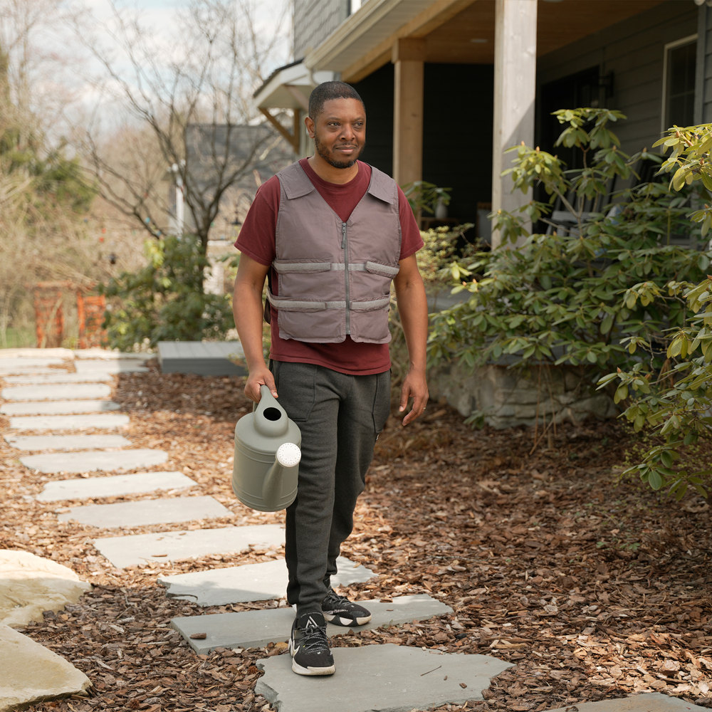 Man walking outdoors holding a watering can, wearing a gray adjustable cooling vest and maroon shirt.