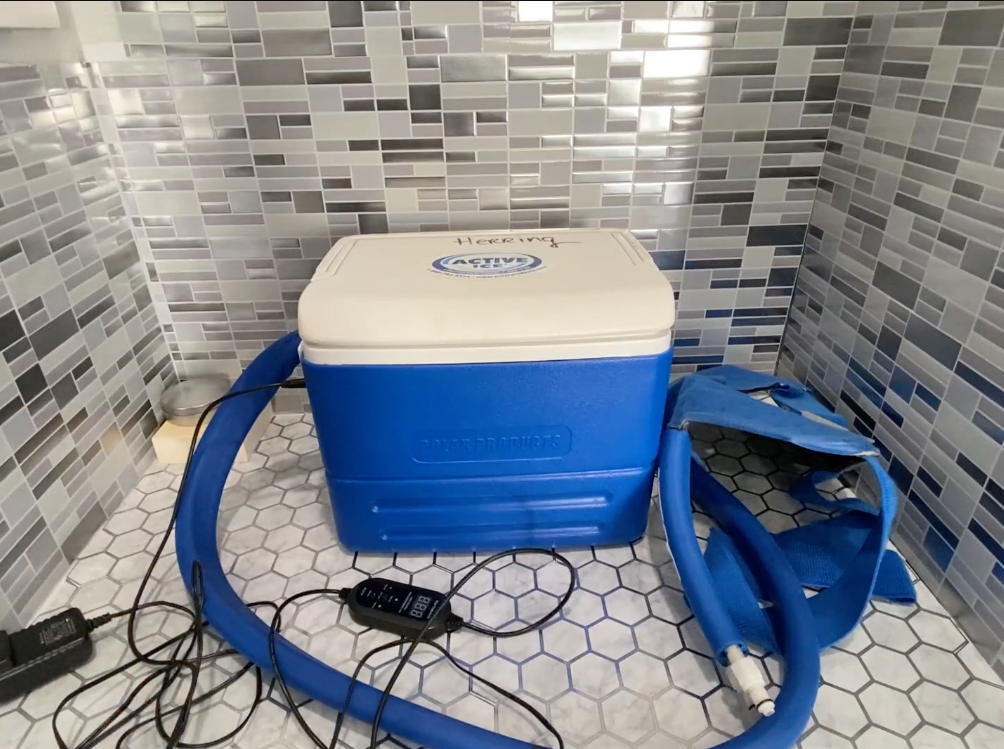 A blue and white Active Ice® Broad Shoulder System with attached blue hoses and a power cord, placed on a tiled floor against a mosaic tile wall.