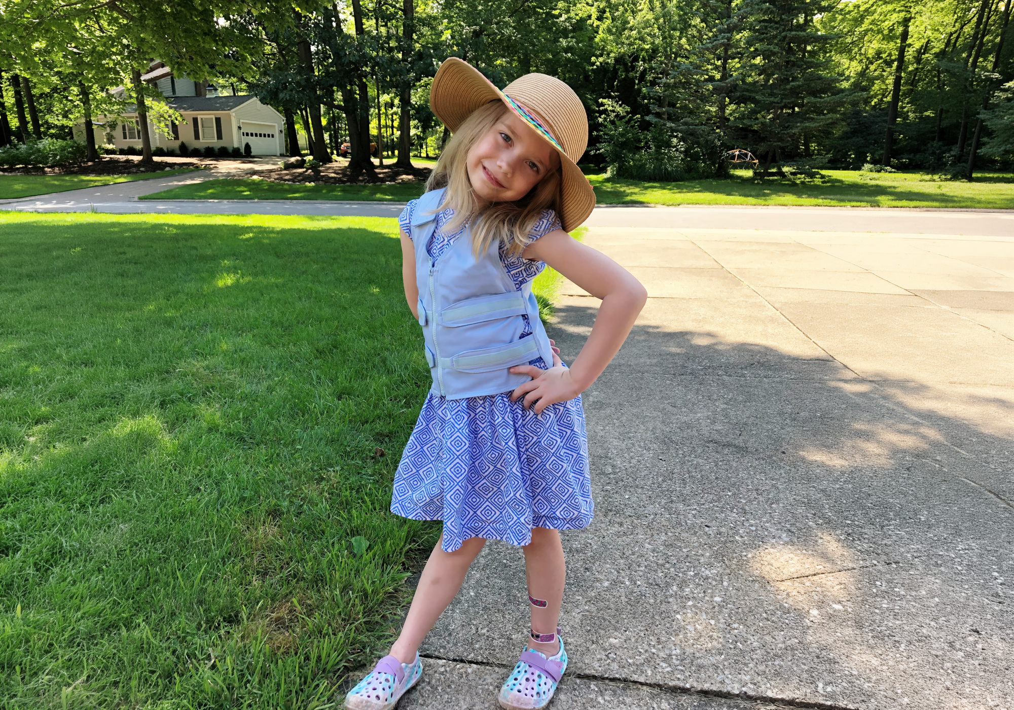Young girl in a blue dress and light blue adjustable cooling vest and straw hat standing on a sidewalk with grass and trees in the background