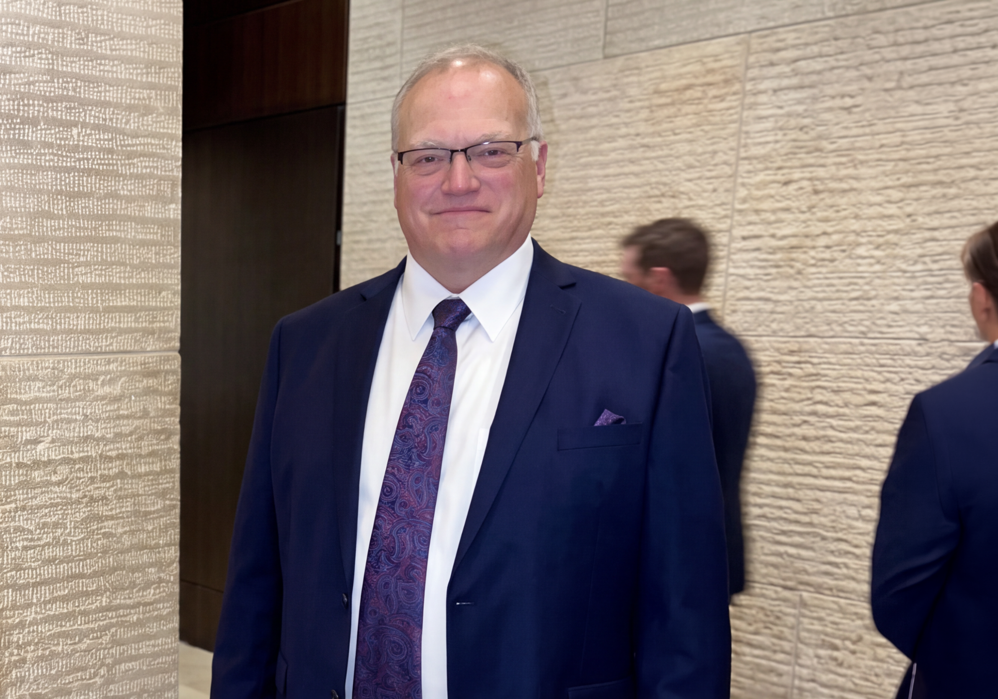 A man wearing a navy blue suit, white dress shirt, and a purple patterned tie, standing in front of a textured beige stone wall.
