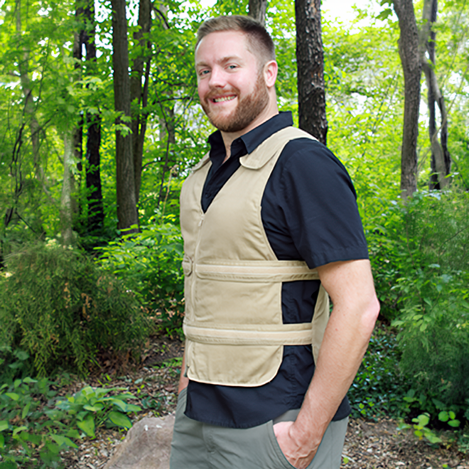 A smiling man wearing a tan adjustable cooling vest over a black short-sleeve shirt stands outdoors in a wooded area with trees and greenery in the background.