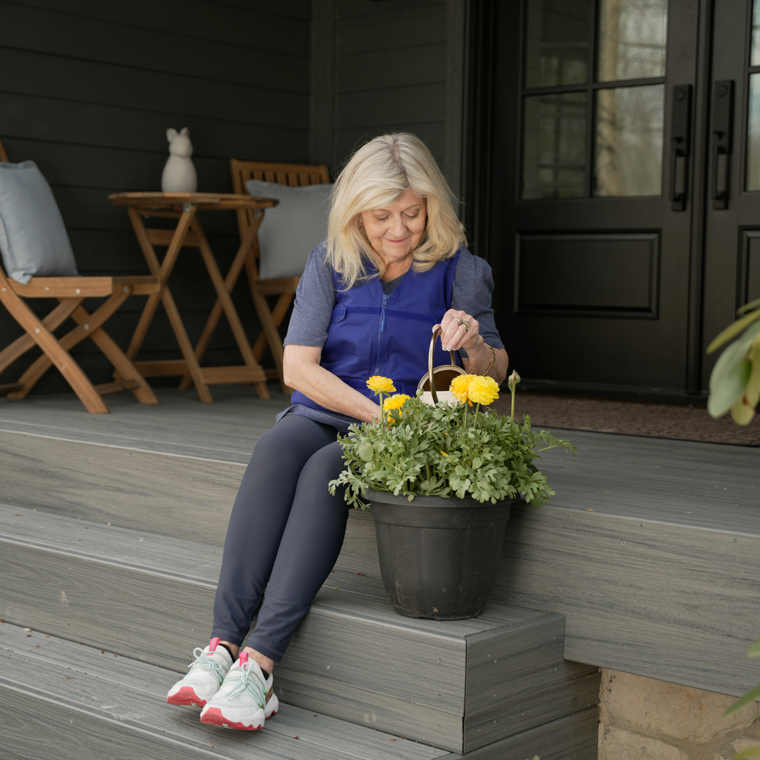 A woman sits on wooden porch steps beside a black planter filled with green foliage and bright yellow flowers, wearing a blue adjustable cooling vest and gray leggings in front of a dark house porch.