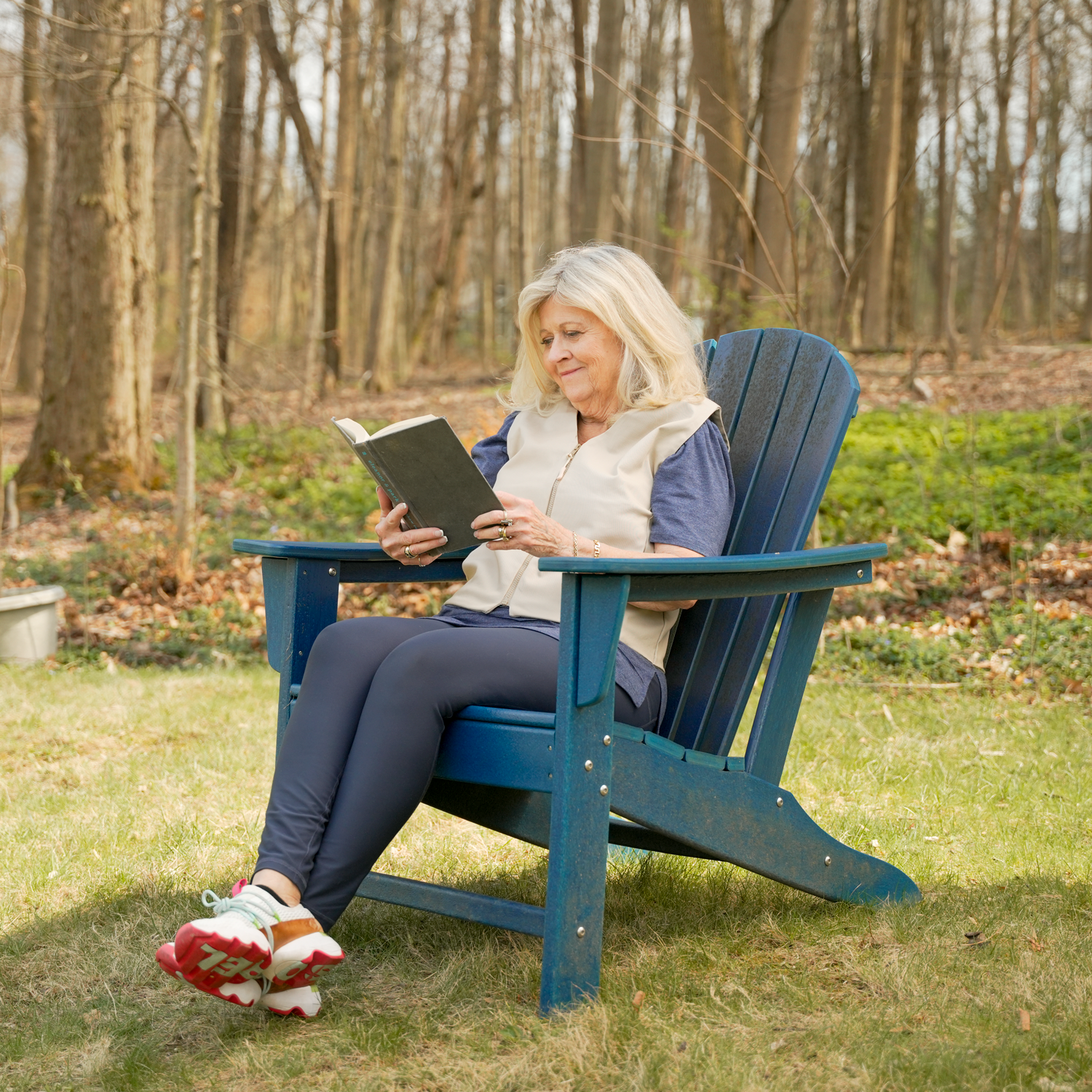 An older woman sits in a blue Adirondack-style outdoor chair on grass, reading a book in a wooded yard wearing a khaki fashion cooling vest. 