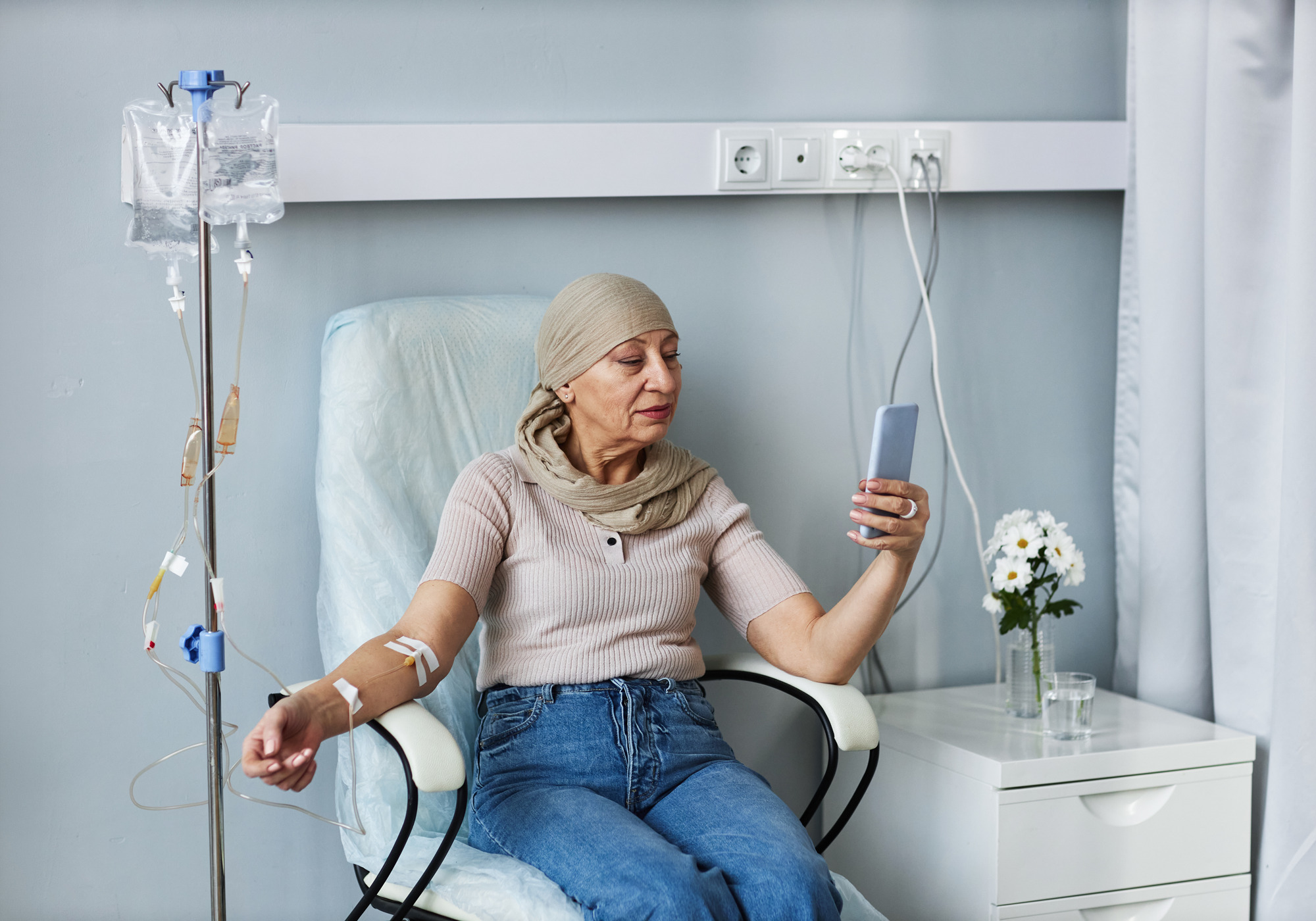 Woman receiving chemotherapy in a hospital room, using a smartphone.