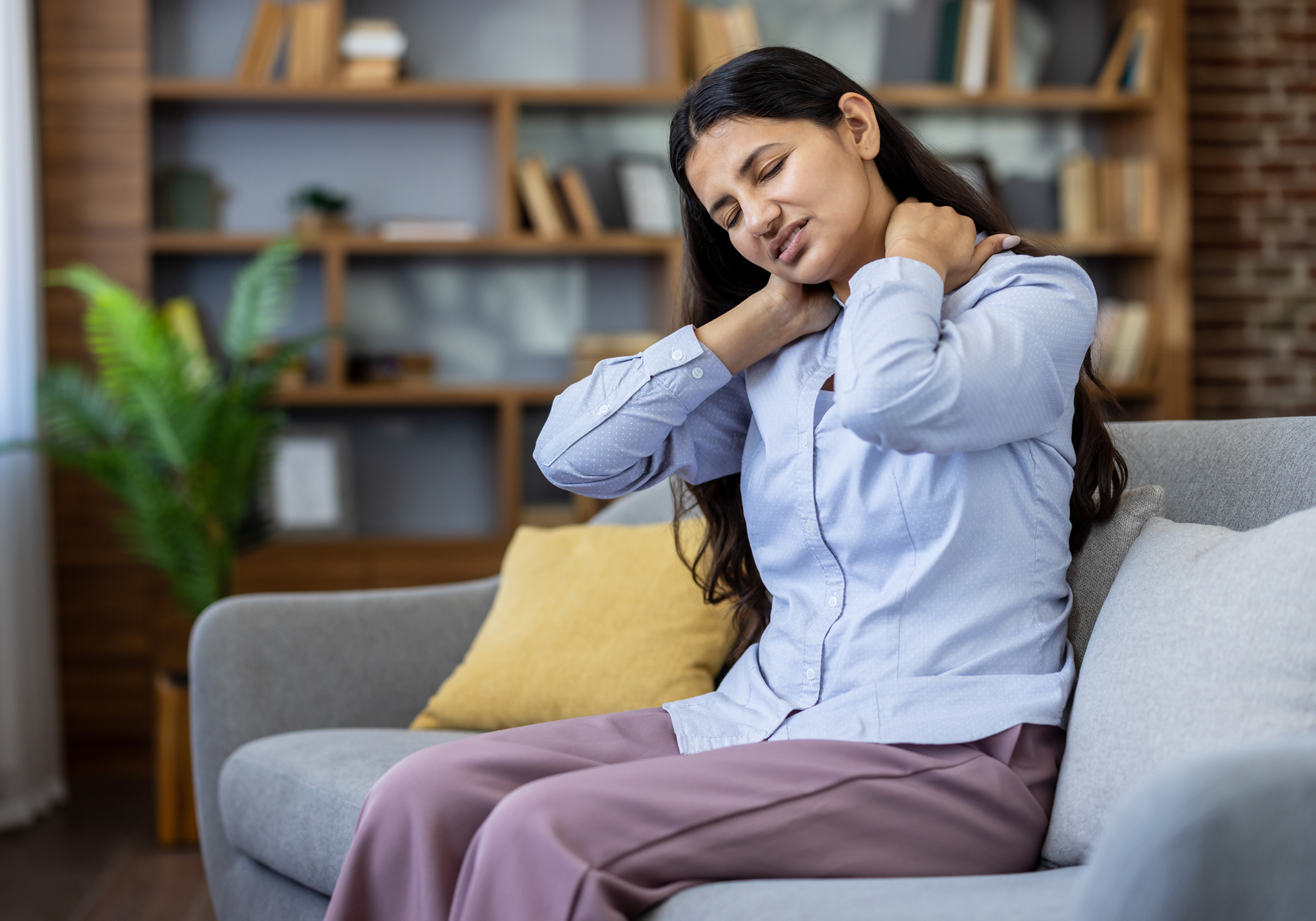 Woman sitting on a couch in a living room, holding her neck and shoulders experiencing neck pain.