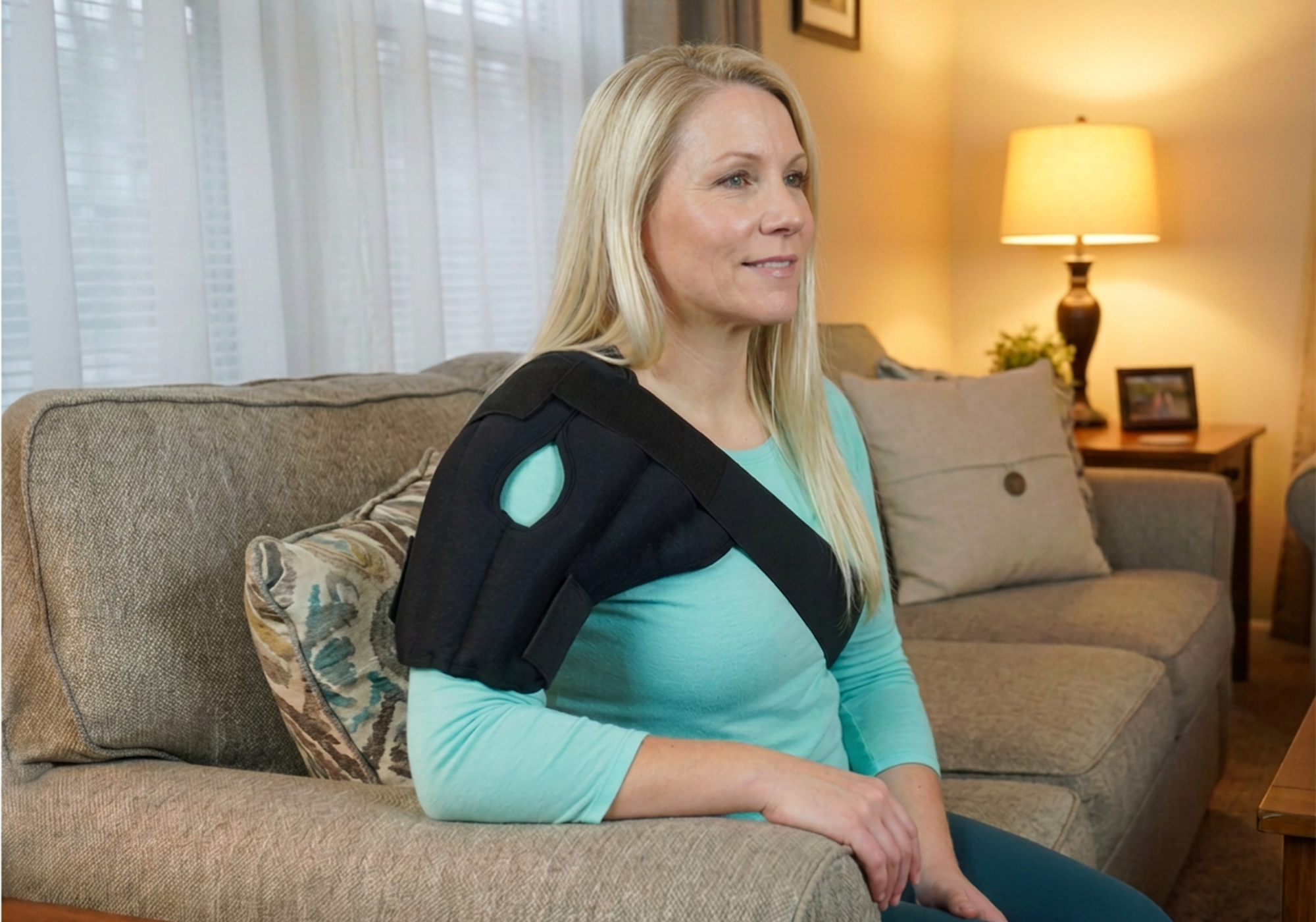 A woman sitting on a couch wearing a black shoulder thera-temp® moist heat wrap over her right shoulder in a warmly lit living room.